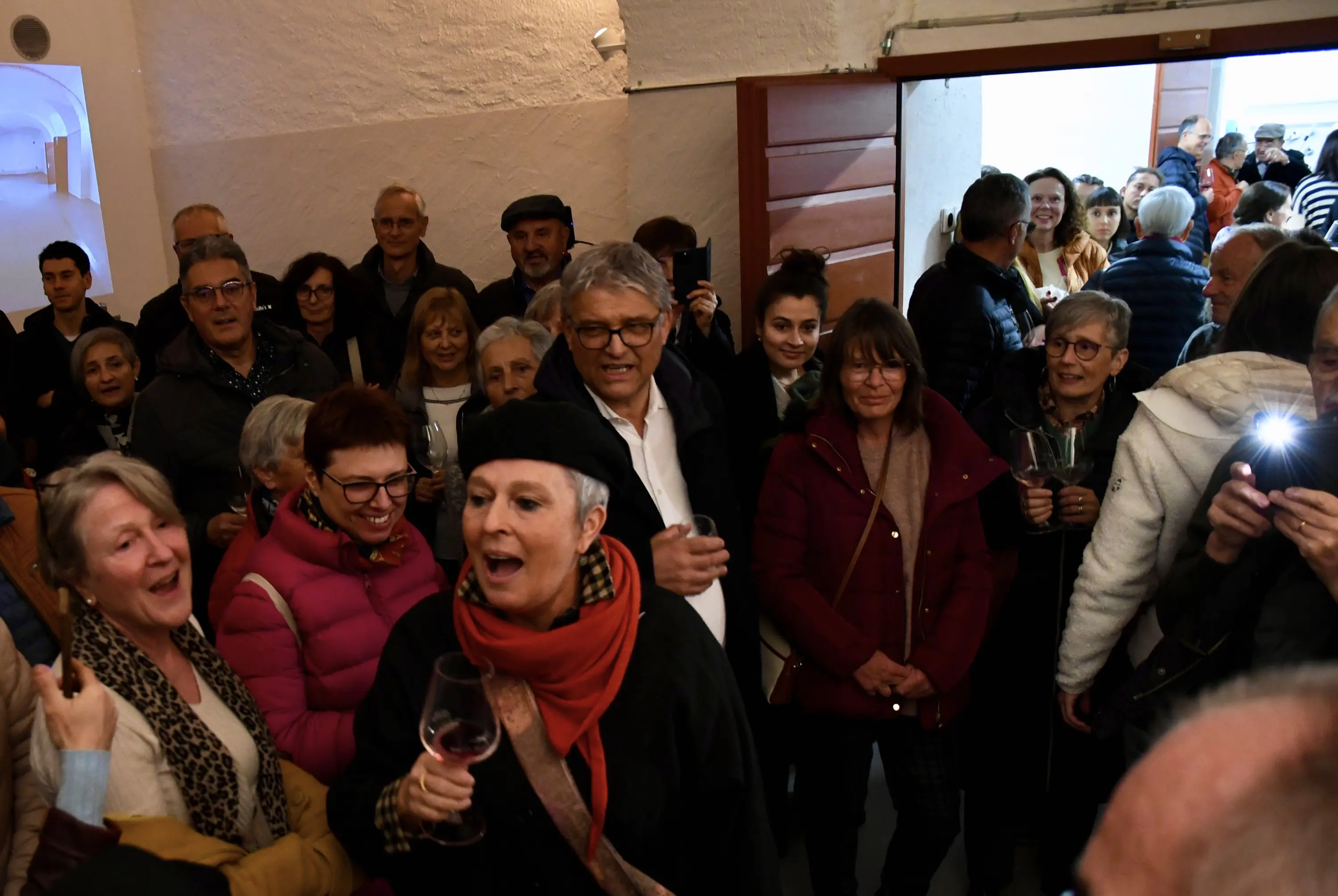 Una folla di persone in un ambiente festoso, alcune con bicchieri di vino in mano, sorridono e chiacchierano. Alcuni atteggiamenti vivaci e molte espressioni di gioia.