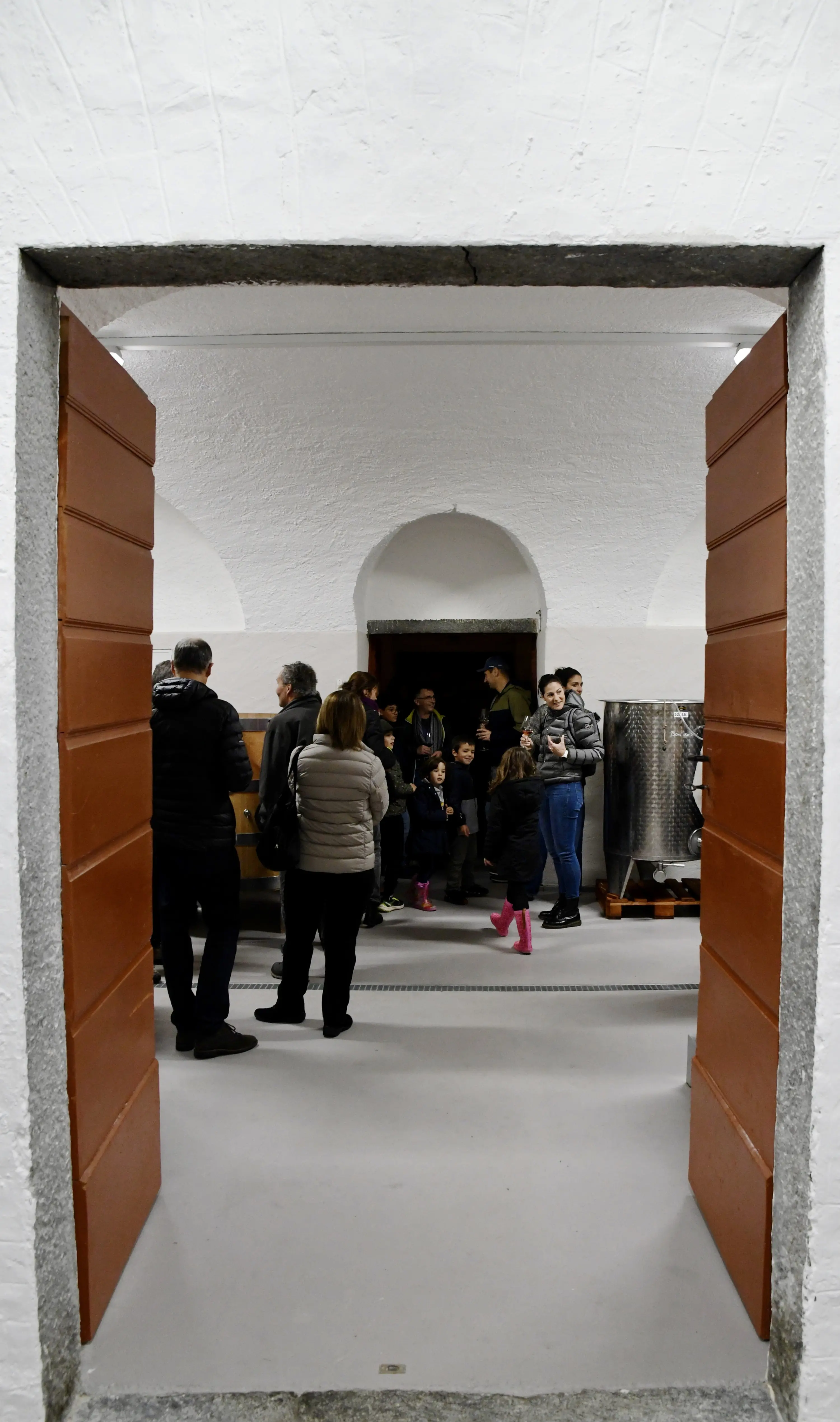 Un gruppo di persone si raduna all'interno di una cantina, con alcuni adulti e bambini visibili. La scena è vista attraverso una porta di legno aperta, mostrando un ambiente semplice e accogliente.