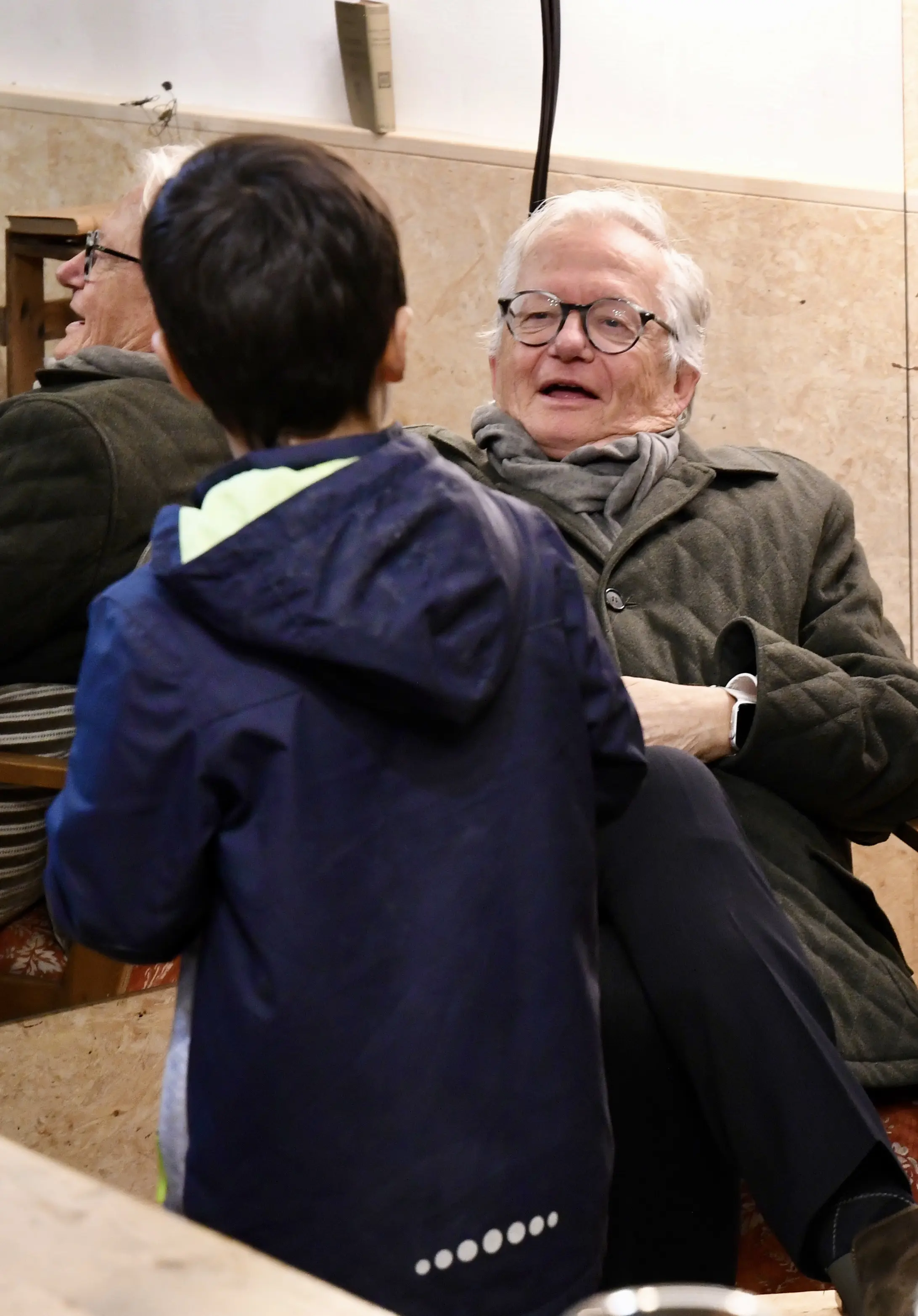 Un uomo anziano con occhiali e sciarpa sorride mentre parla con un bambino. Il bambino si volta verso di lui, indossando un giubbotto blu. Sullo sfondo si vede un riflesso nell’acqua.