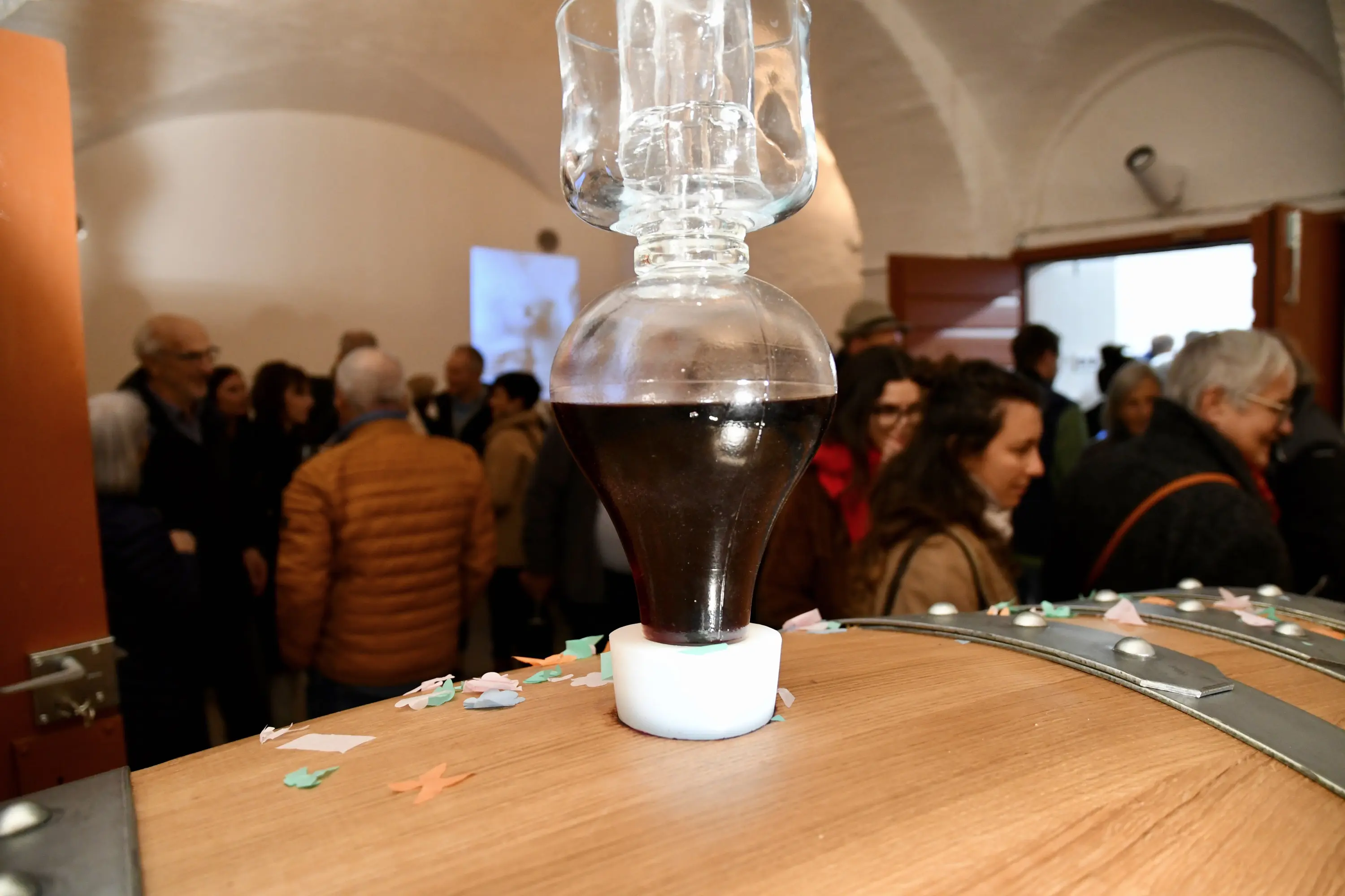 Un contenitore di vetro con liquido scuro, appoggiato su un barile di legno, in primo piano. Sullo sfondo, un gruppo di persone conversano in un ambiente luminoso e accogliente. Decorazioni colorate e festoni sparsi sul tavolo