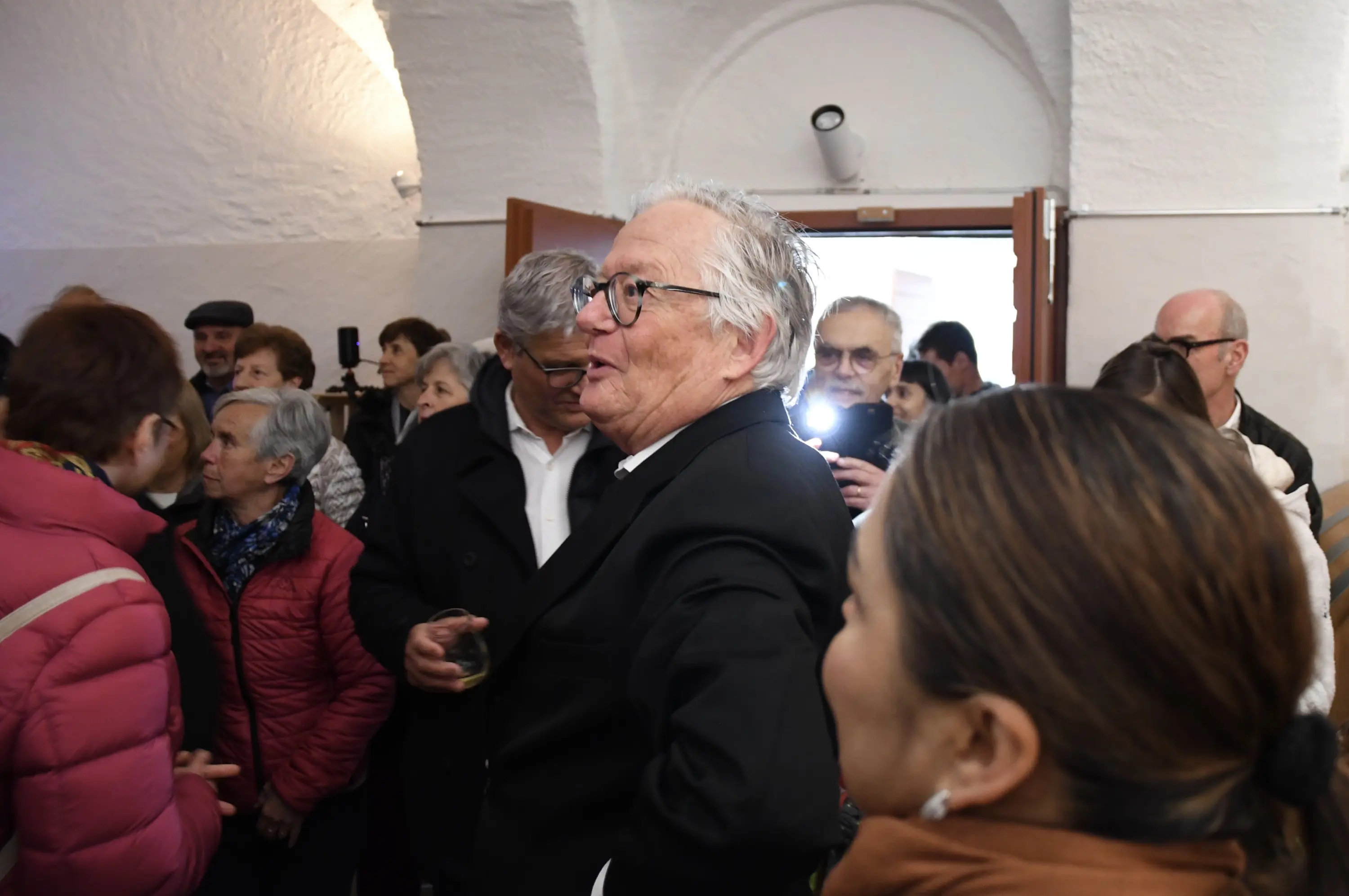 Un uomo con occhiali e capelli grigi parla a un gruppo di persone in un ambiente interno affollato, mentre alcuni partecipanti sorridono e lo ascoltano attentamente.