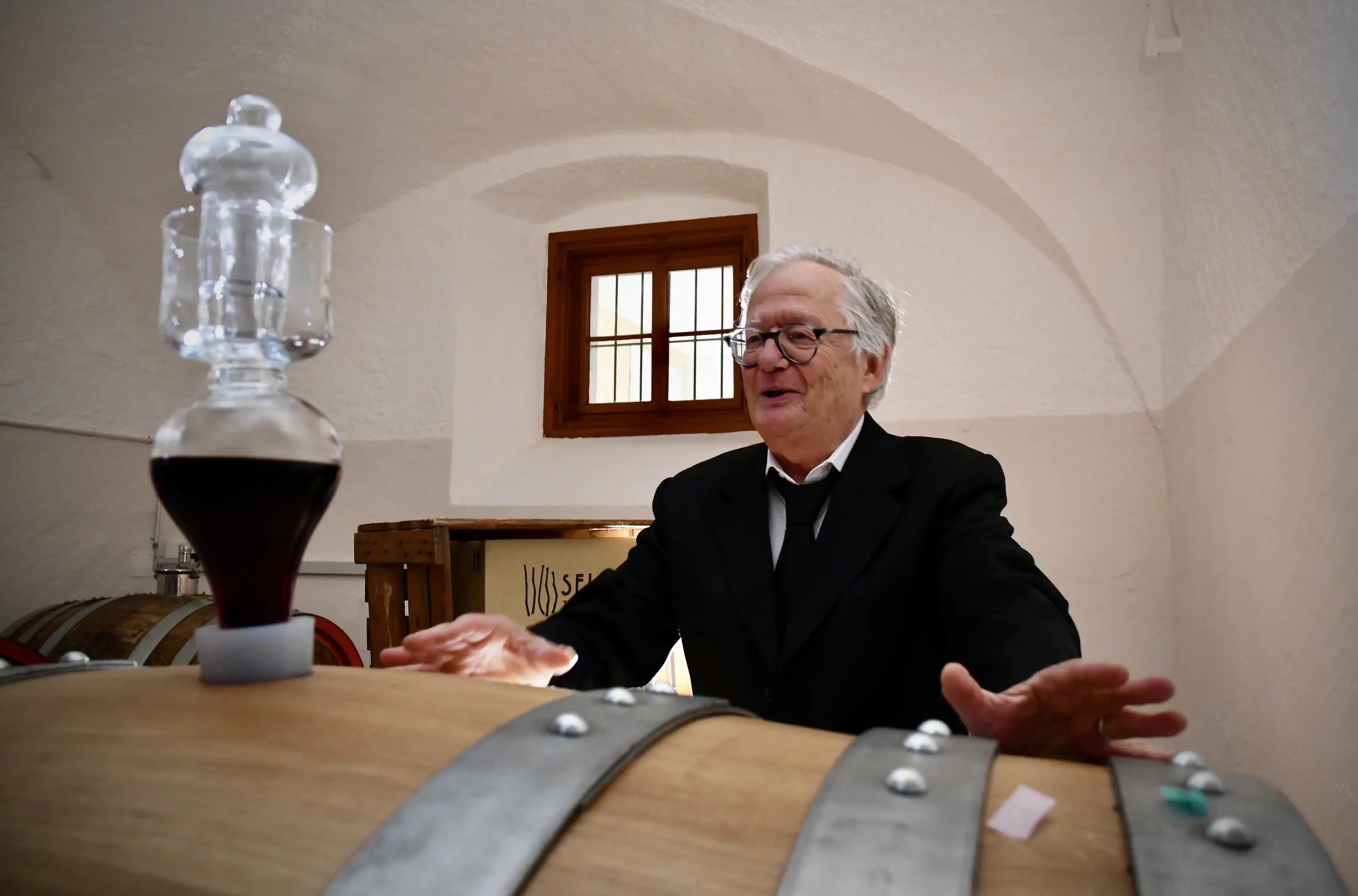 Un uomo anziano, vestito elegantemente, si trova davanti a una grande botte di legno. Sorride mentre osserva un contenitore di vetro con una sostanza scura sopra la botte. Sullo sfondo, si intravede una finestra e alcune botti