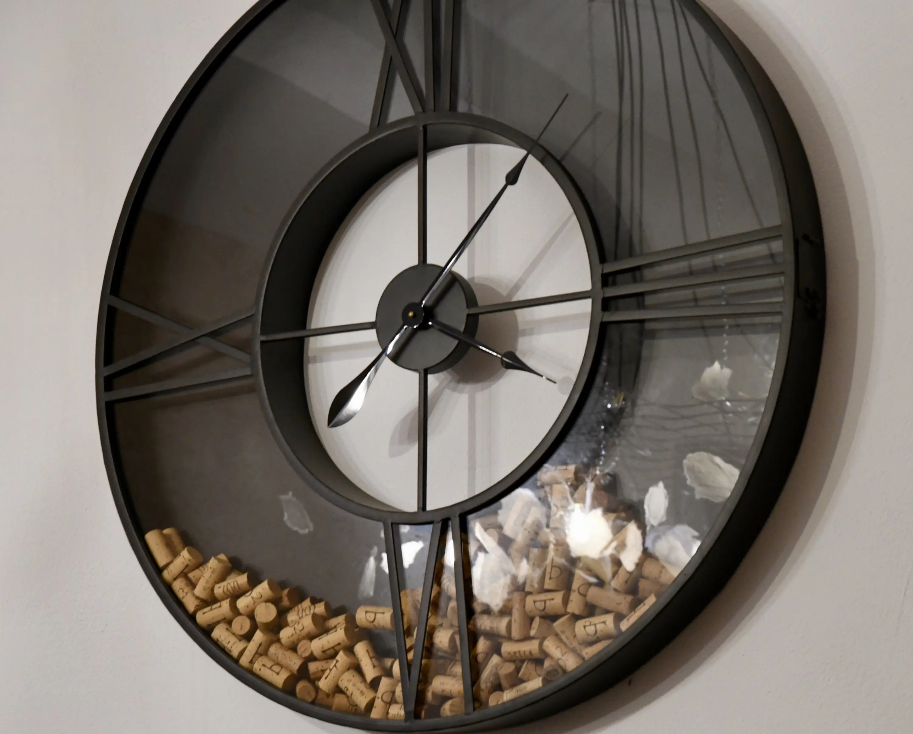 Orologio da parete rotondo con cornice nera, trasparente al centro e riempito con tappi di sughero sul fondo. Le lancette dell'orologio sono visibili attraverso il vetro.