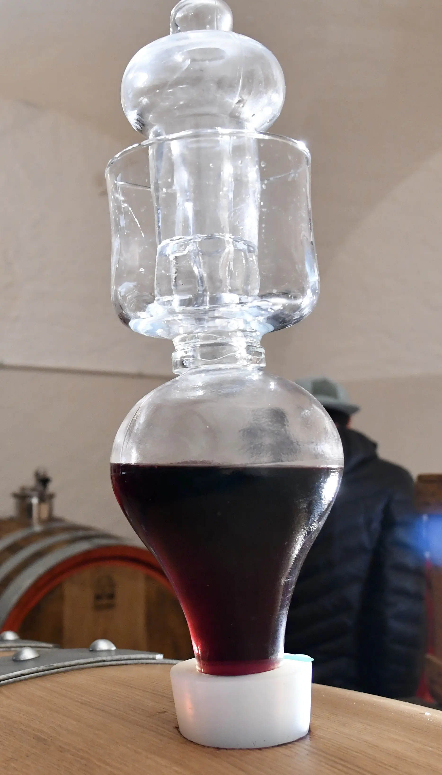 Un dispositivo di vetro per la decantazione del vino, con un liquido rosso all'interno. Sullo sfondo si intravedono alcune botti di legno.