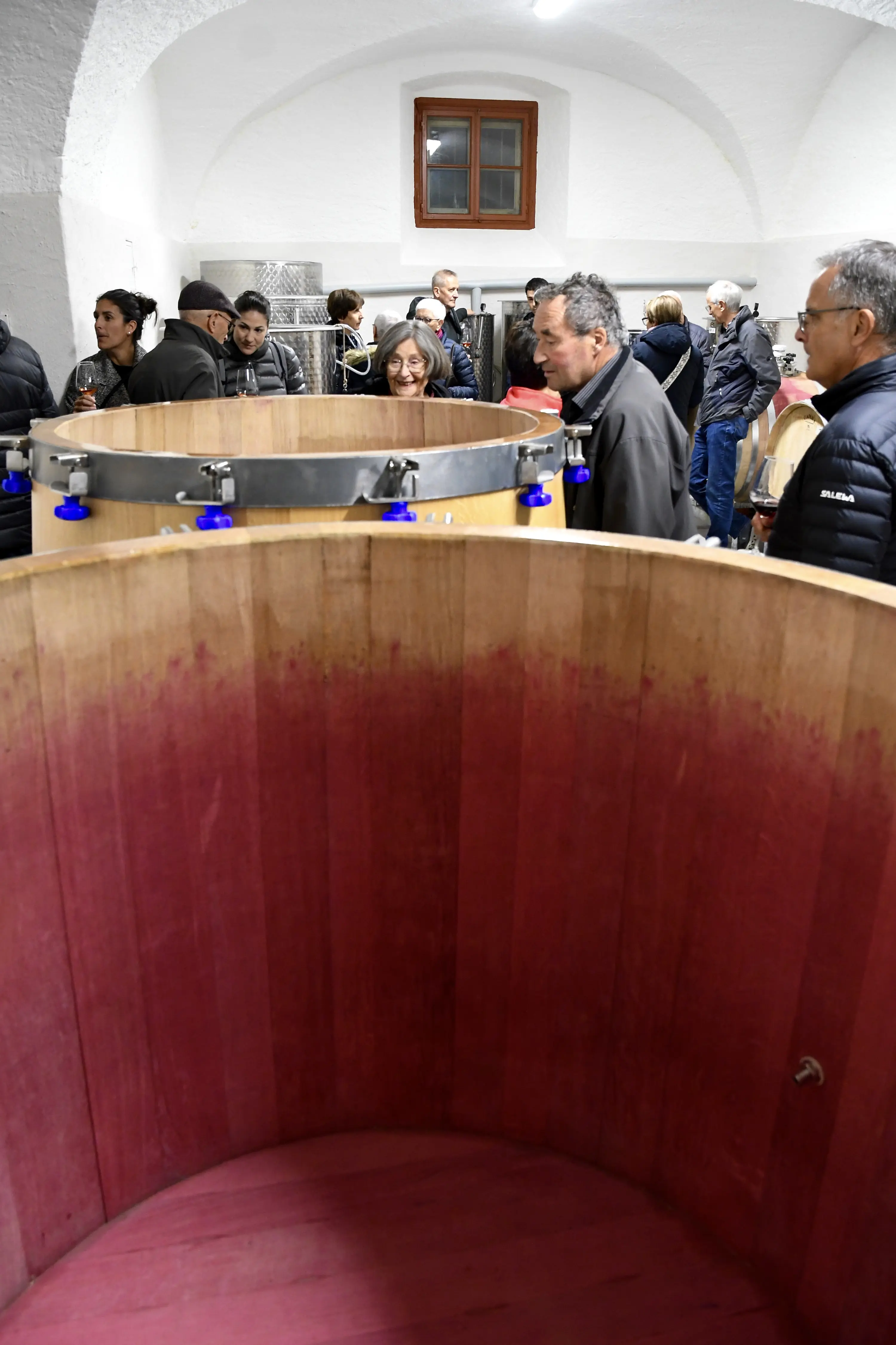Un gruppo di persone si riunisce in una cantina, con un grande recipiente di legno in primo piano. Alcuni individui conversano, mentre altri osservano l'ambiente circostante. Gli elementi tipici della degustazione di vini si fanno notare.
