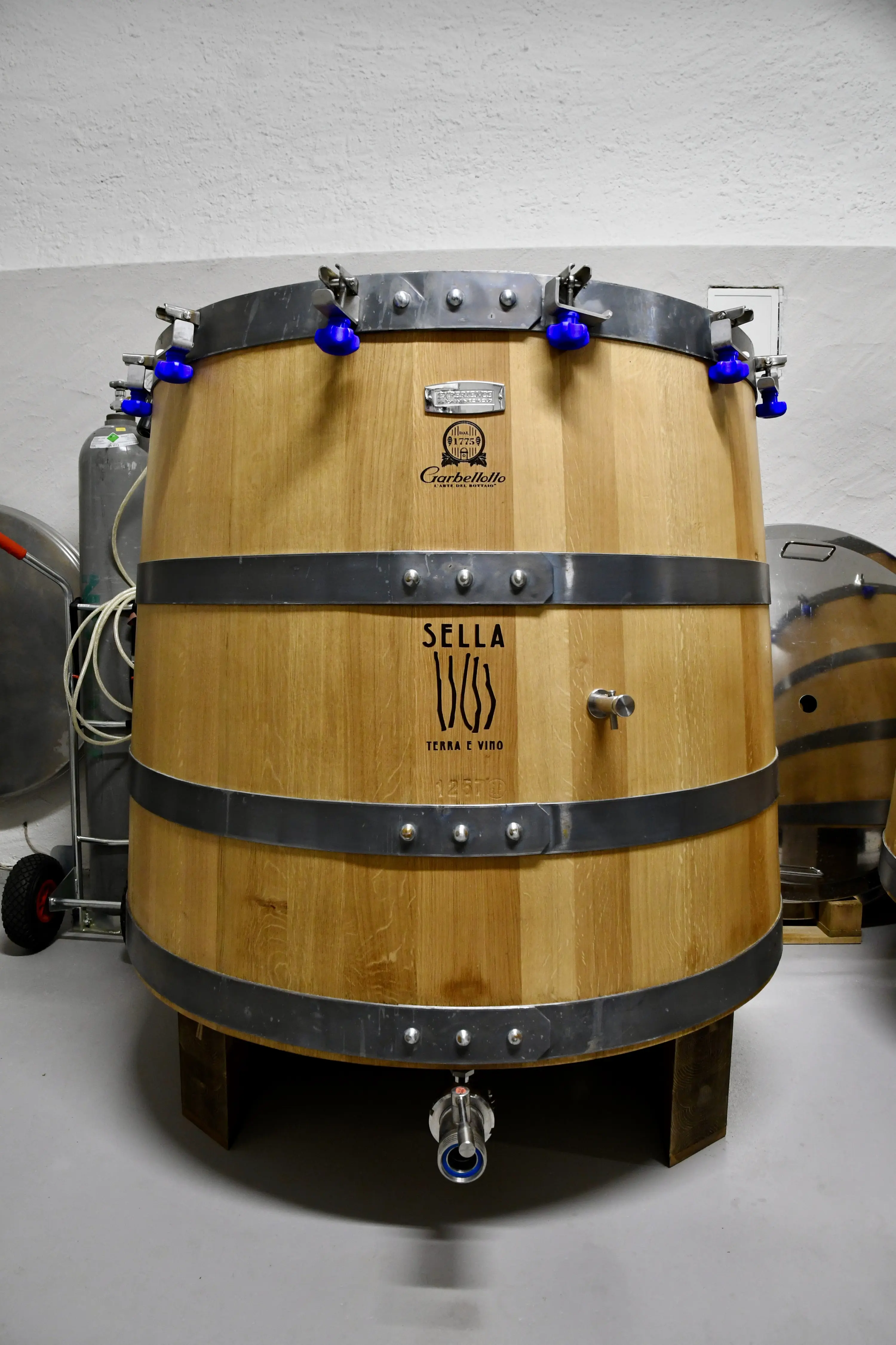 Un grande tino di legno per la fermentazione del vino, con bande metalliche e rubinetto, situato in un ambiente di cantina.