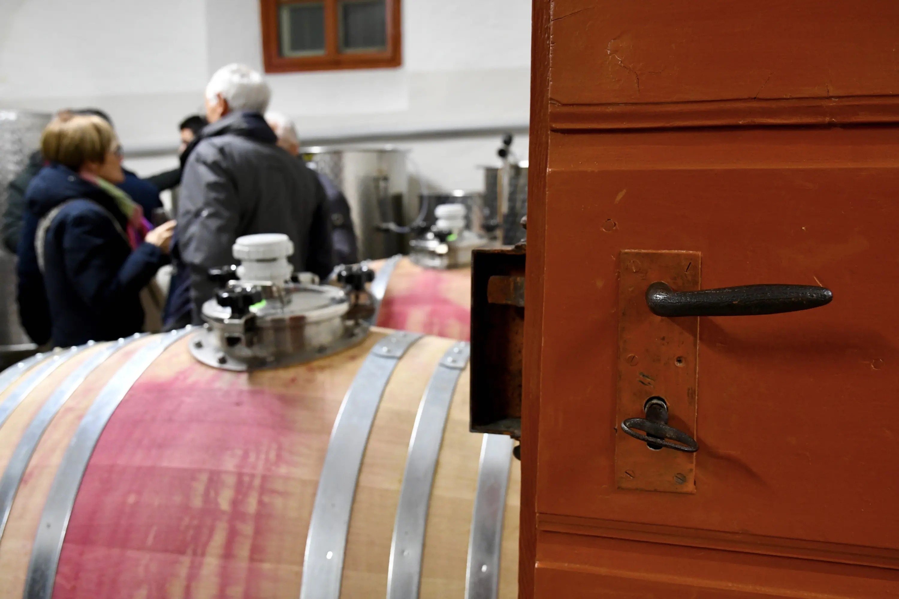 Una porta di legno con una maniglia in metallo, affiancata da una grande botte di legno. Sullo sfondo, alcune persone stanno conversando in un ambiente di cantina.