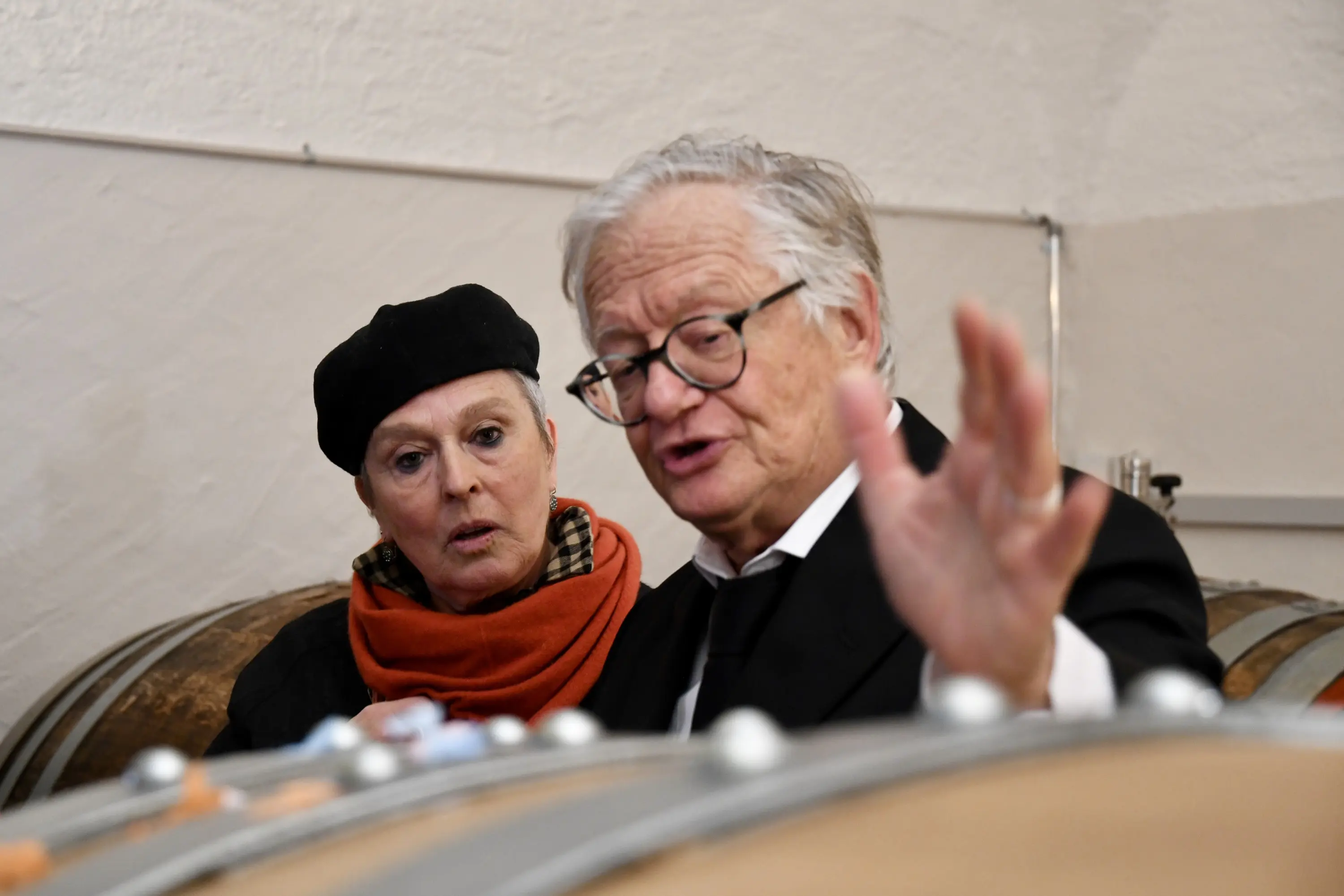 Due persone in un ambiente di cantina vinicola, una donna con un berretto e una sciarpa arancione osserva attentamente, mentre un uomo in abito scuro spiega qualcosa, gesticolando verso una botte.