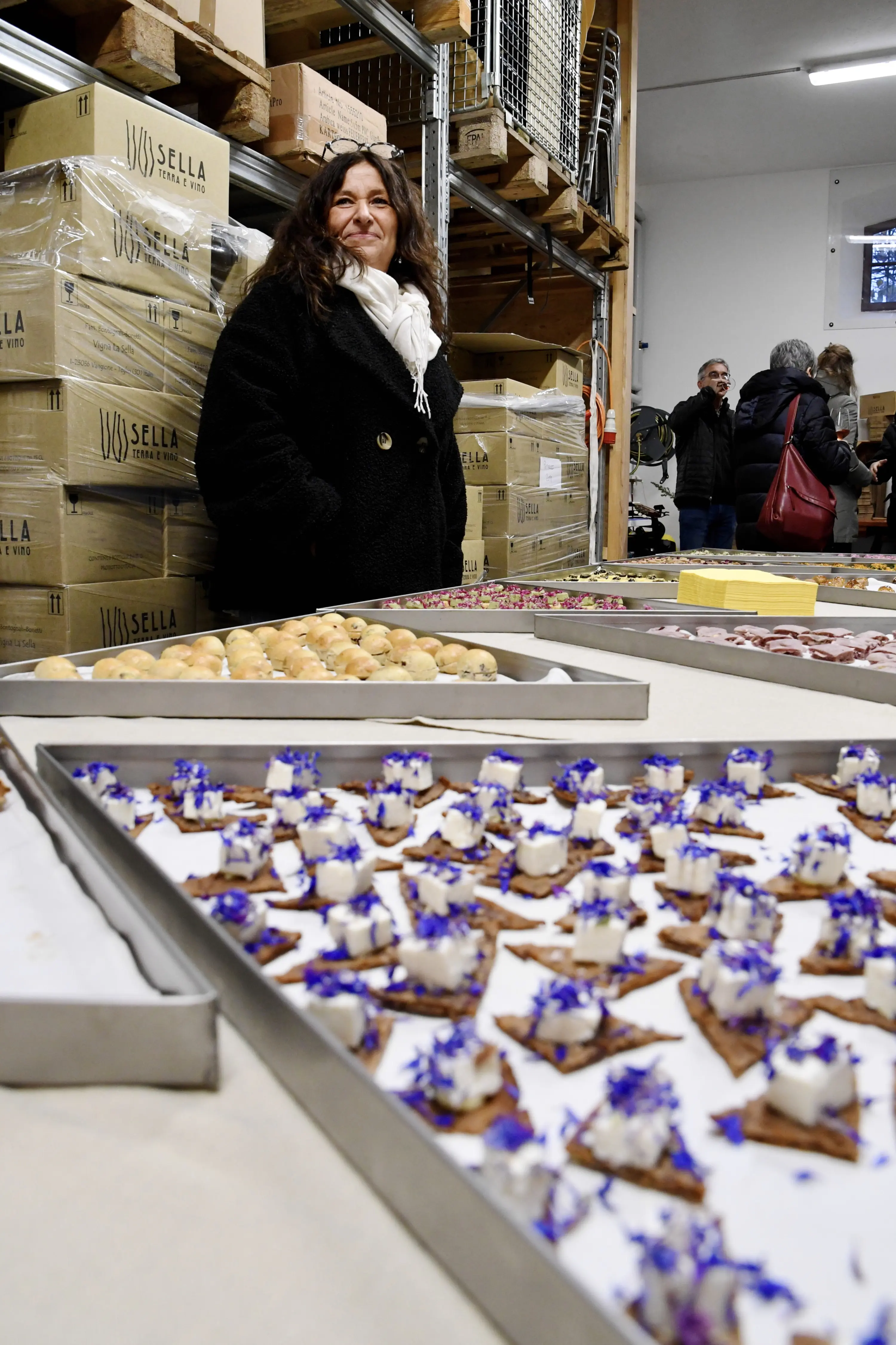 Una donna sorridente in un laboratorio, accanto a piatti di dolci decorati con fiori blu. Sullo sfondo, persone che chiacchierano e scatole di prodotti in legno.