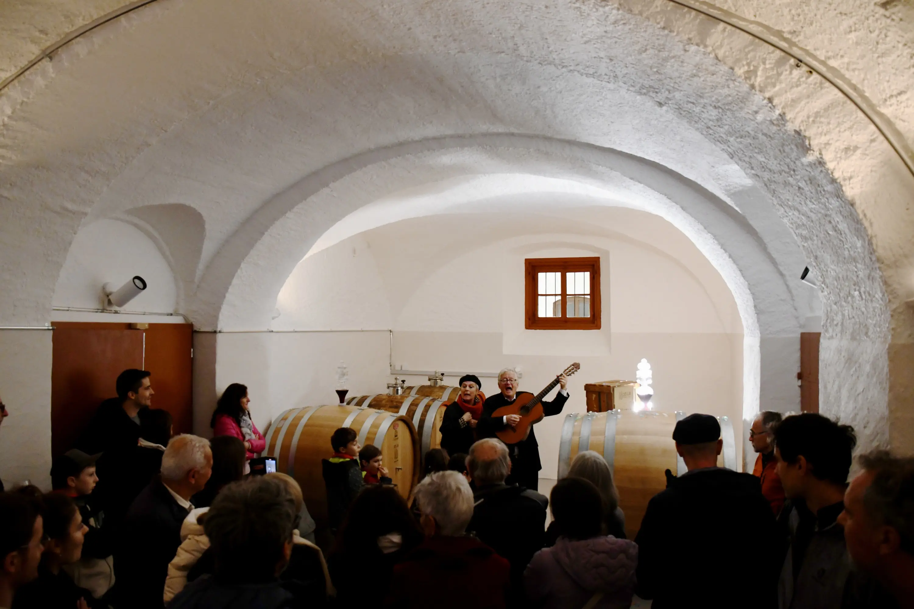 Una cantante si esibisce con una chitarra in una cantina, circondata da un gruppo di persone. Sullo sfondo si vedono grandi botti di legno e un soffitto a volta.