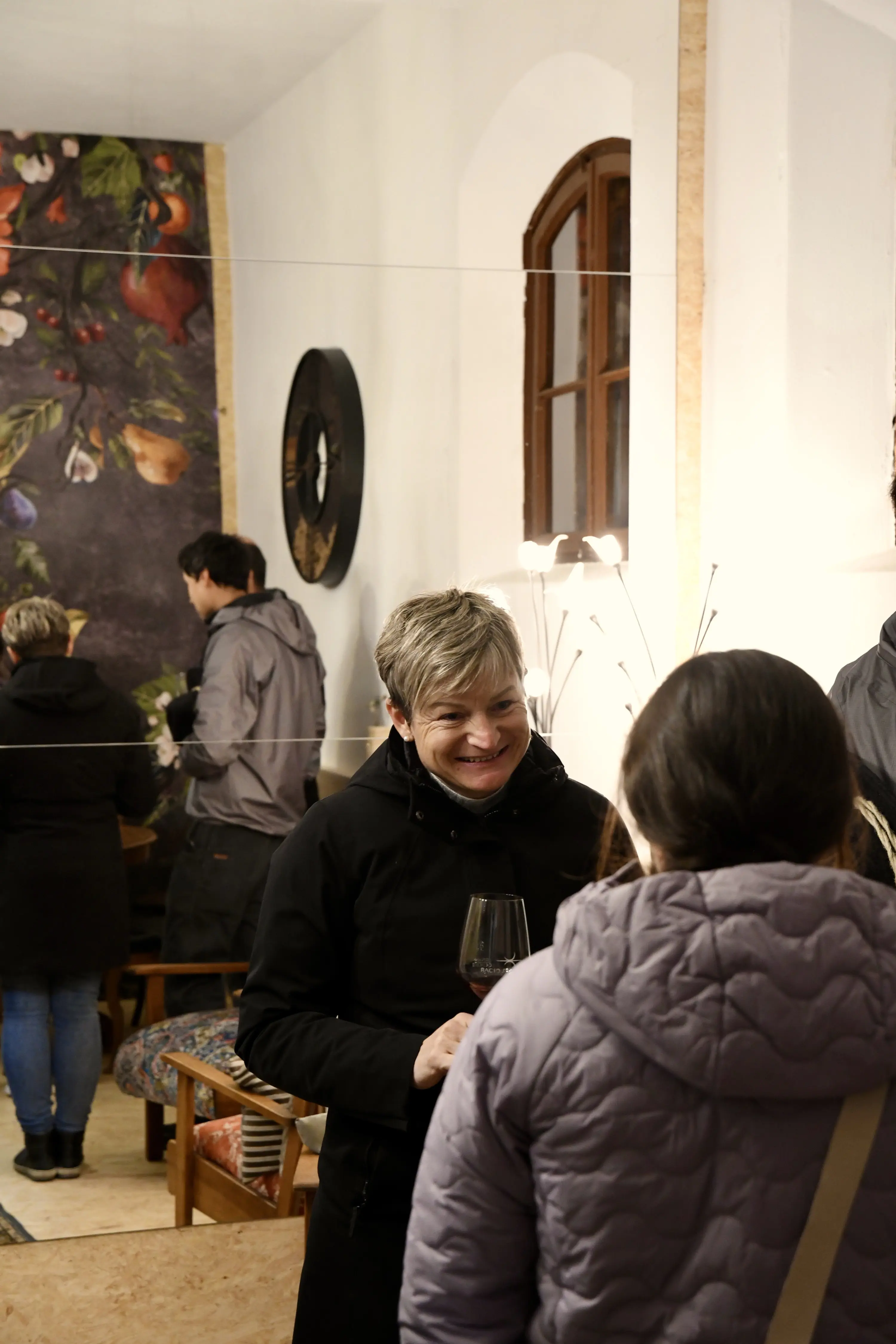 Una donna sorridente conversa con un'altra persona in un ambiente accogliente, con pareti decorate e un tavolo in legno. Altre persone sono visibili sullo sfondo, creando un'atmosfera sociale.