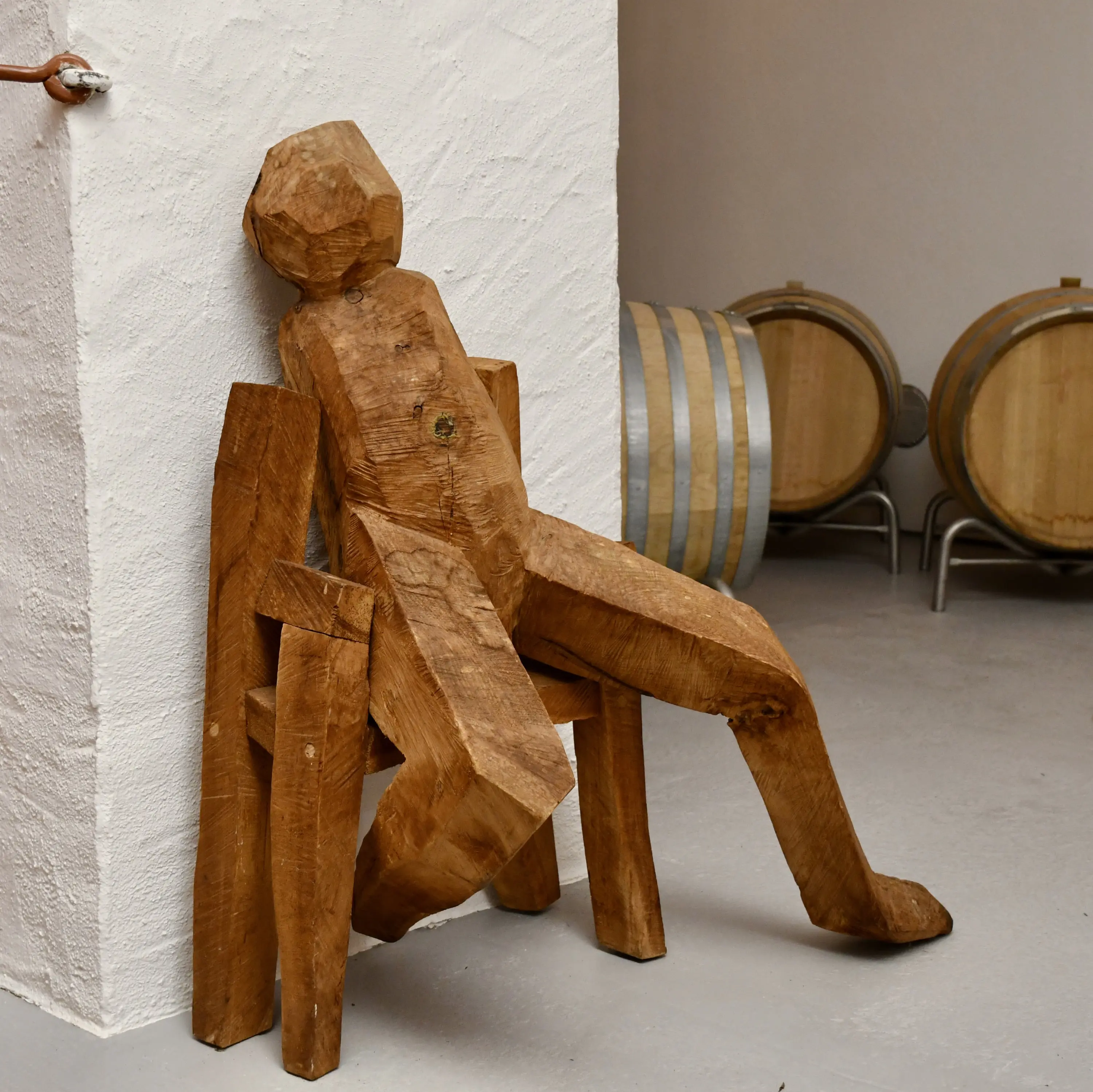 Scultura di legno di una figura stilizzata seduta su una sedia, appoggiata a una parete bianca, con botti di legno sullo sfondo.