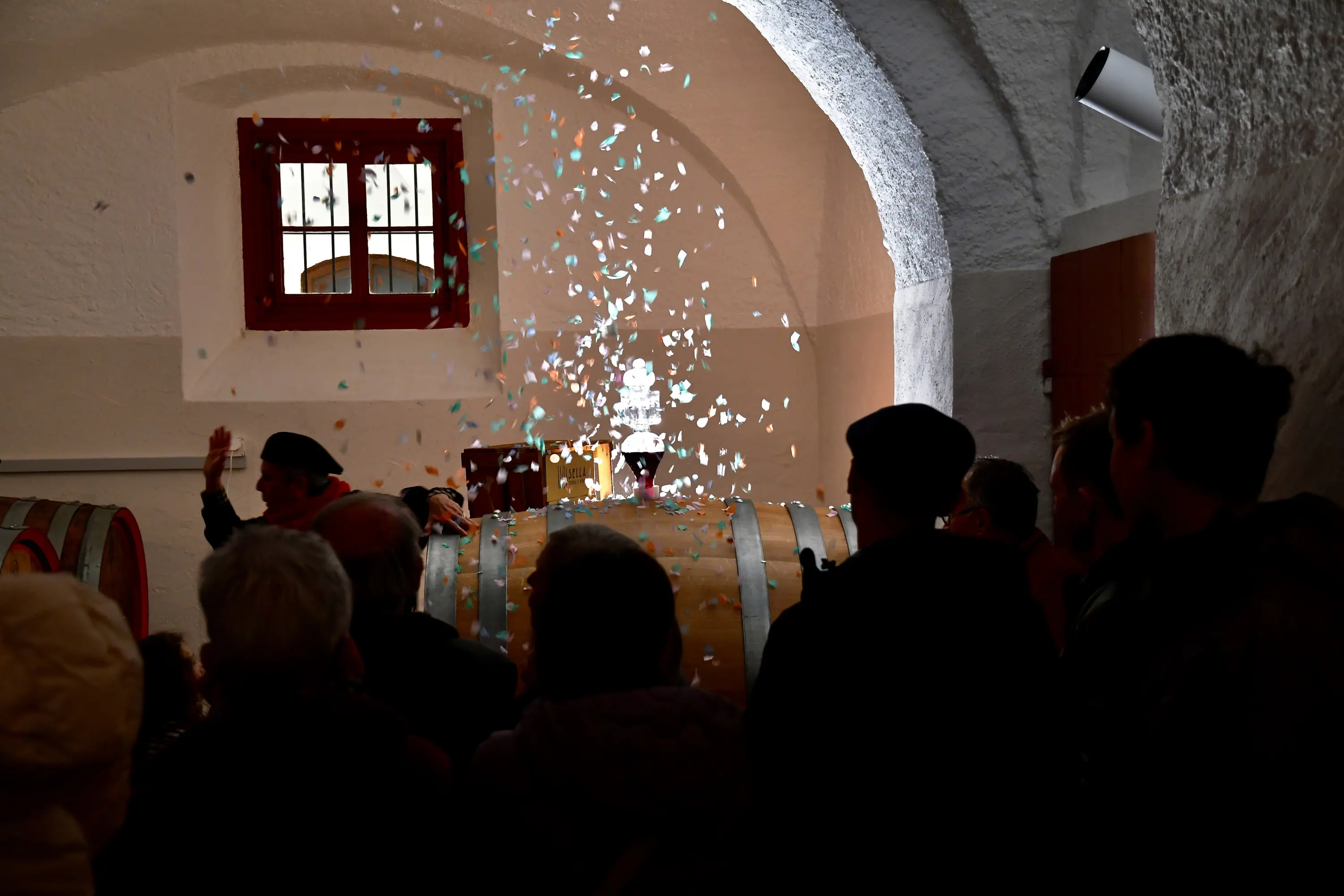 Una folla osserva mentre coriandoli colorati cadono da un alto contenitore in una cantina con botti di legno. La scena è illuminata da luci soffuse e gli spettatori sorridono entusiasti.