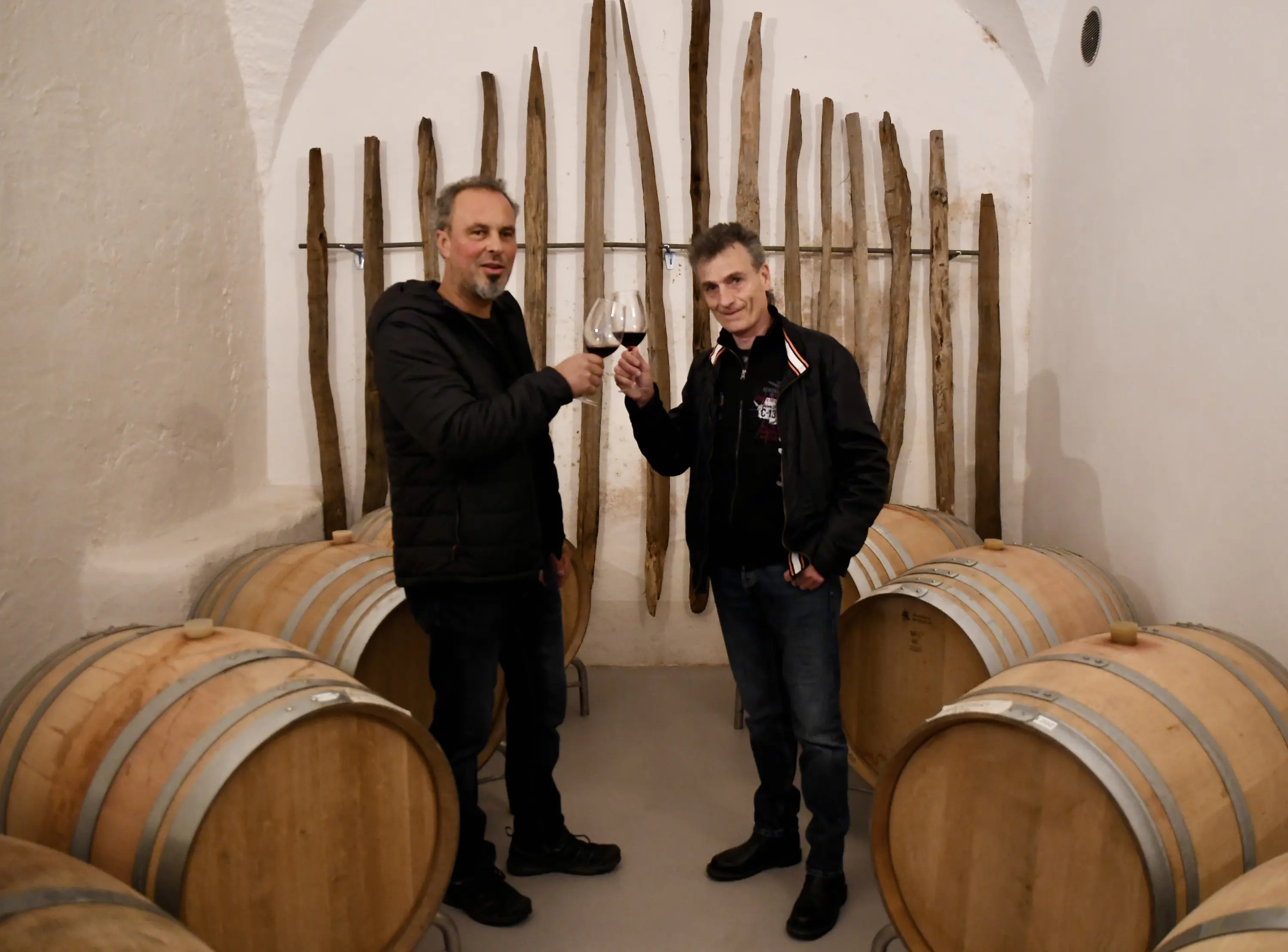 Due uomini sorridenti alzano i bicchieri di vino in una cantina. Sullo sfondo, barili di legno e pareti decorate con doghe di legno.