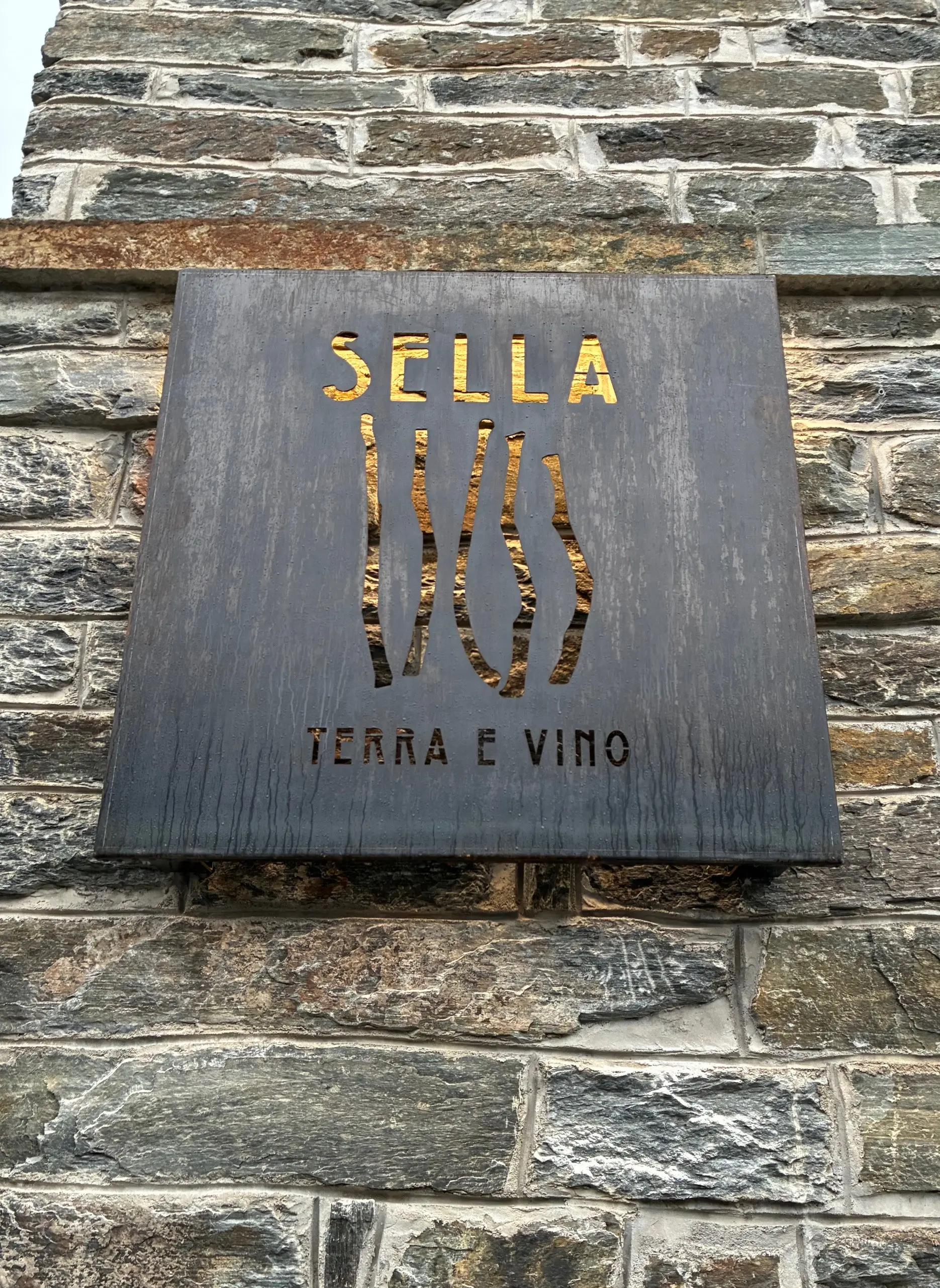 Insegna in legno scuro con il testo "SELLA" in lettere maiuscole dorate, seguito dalla scritta "TERRA E VINO" in caratteri più piccoli. La base è di pietra grigia.