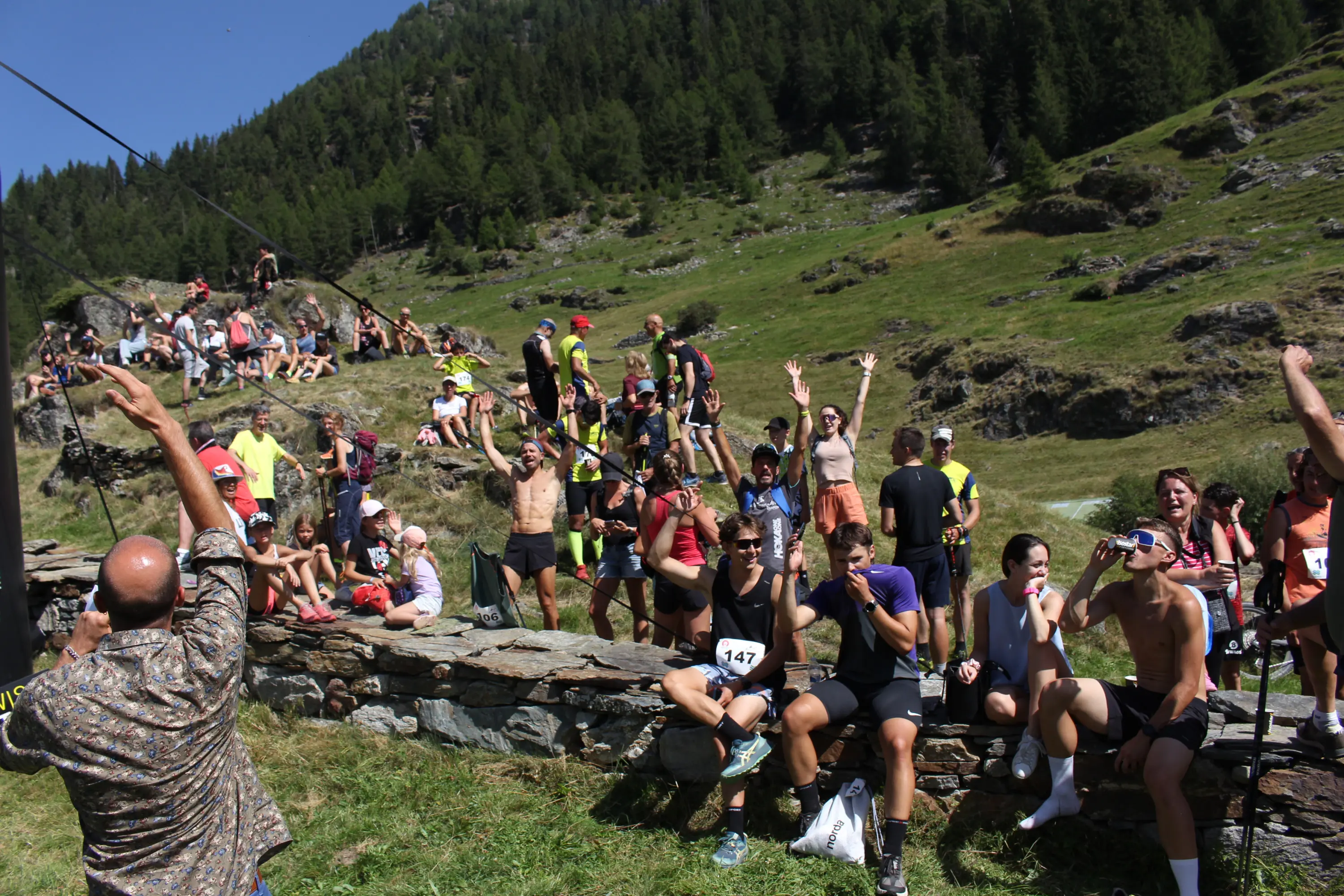Gruppo di persone che partecipano a un evento all'aperto in montagna. Alcuni corridori sono seduti su un muretto, mentre altri sono in piedi e applaudono. Gli spettatori sono vestiti in abbigliamento sportivo e si trovano in un ambiente naturale verde e montuoso.