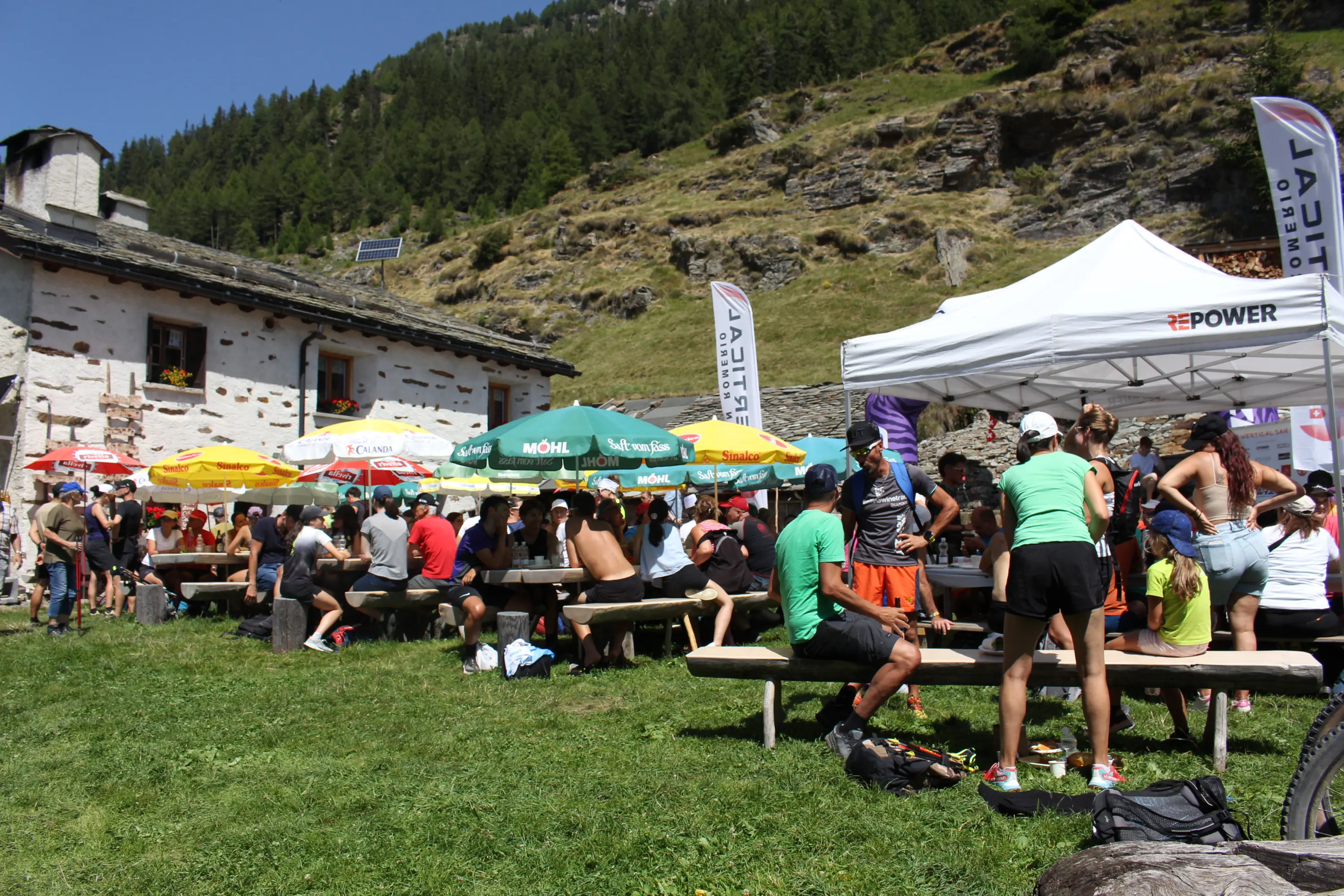 Un gruppo di persone si riposa e socializza all’aperto, seduto su panche di legno in un'area verde. Sullo sfondo si vede un edificio di montagna e tende con ombrelloni colorati. Tra la folla, alcune persone indossano abbigliamento sportivo e si possono notare anche bandiere e striscioni.