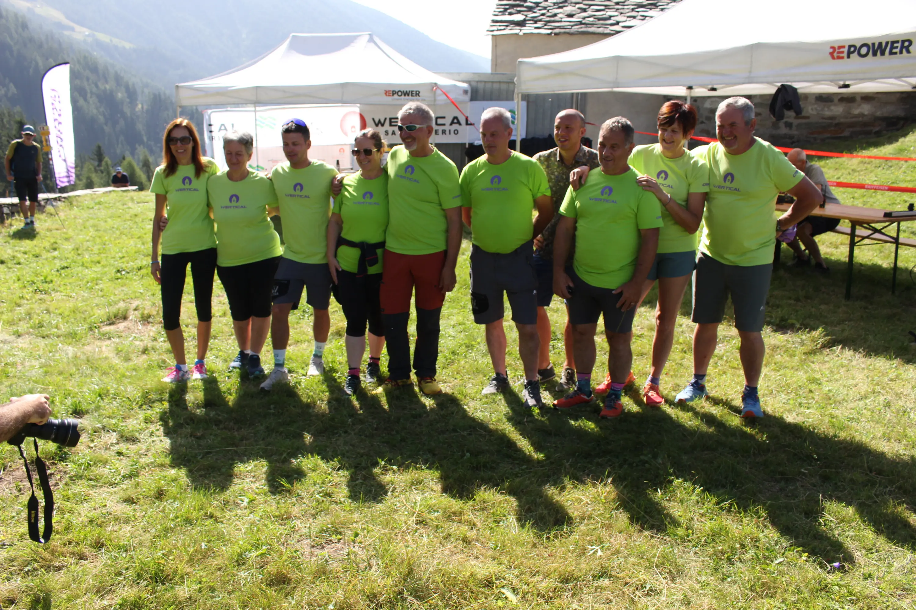 Gruppo di persone in magliette verdi brillanti sorridendo davanti a un gazebo in montagna. Sullo sfondo si vedono alberi e una struttura in legno.