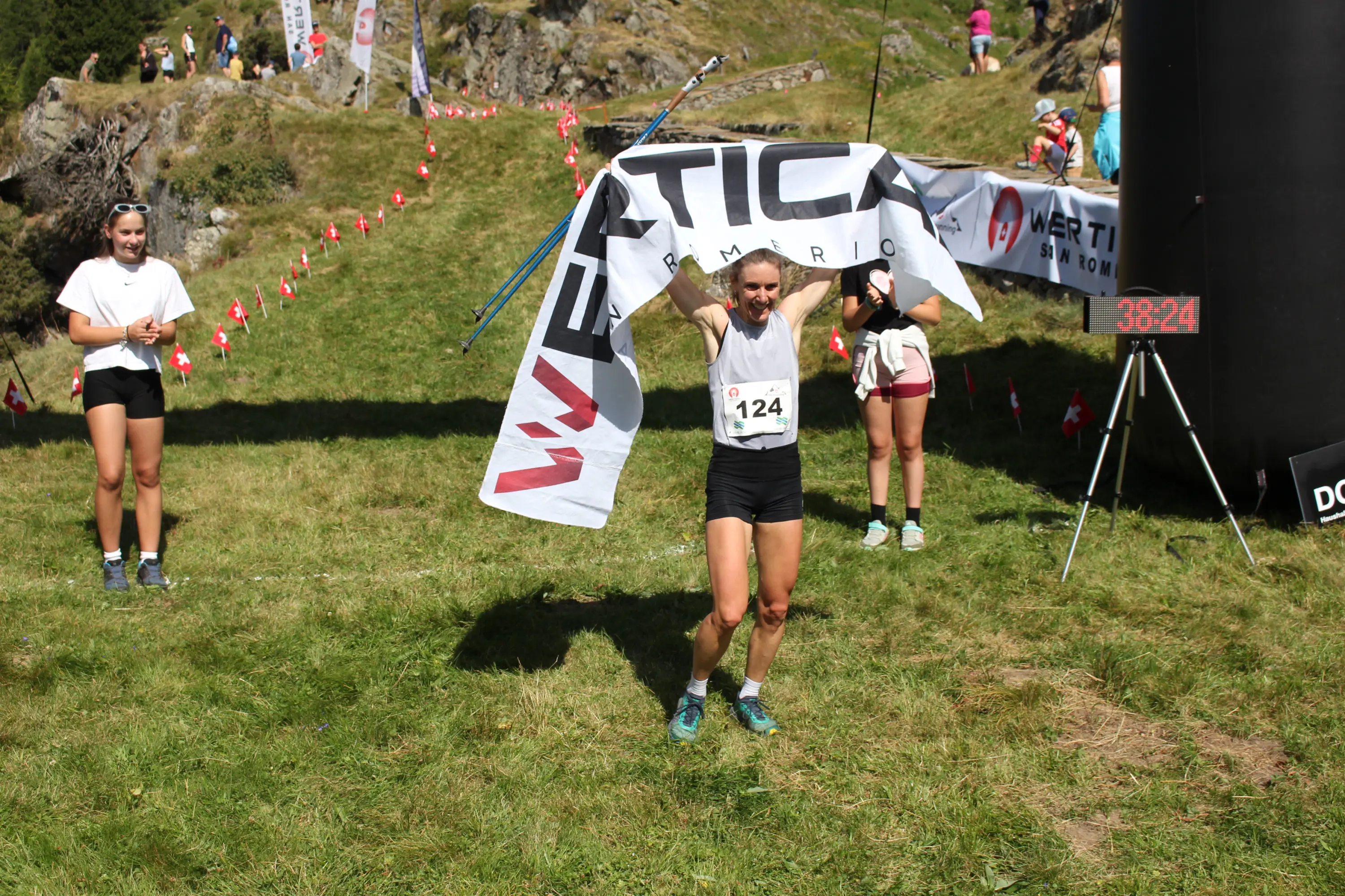 Una corridore attraversa il traguardo sorridente, sollevando una bandiera con il logo di un marchio. In secondo piano, altre due persone applaudono. Un timer segna un tempo di 38:24. Il paesaggio è verde e montano.