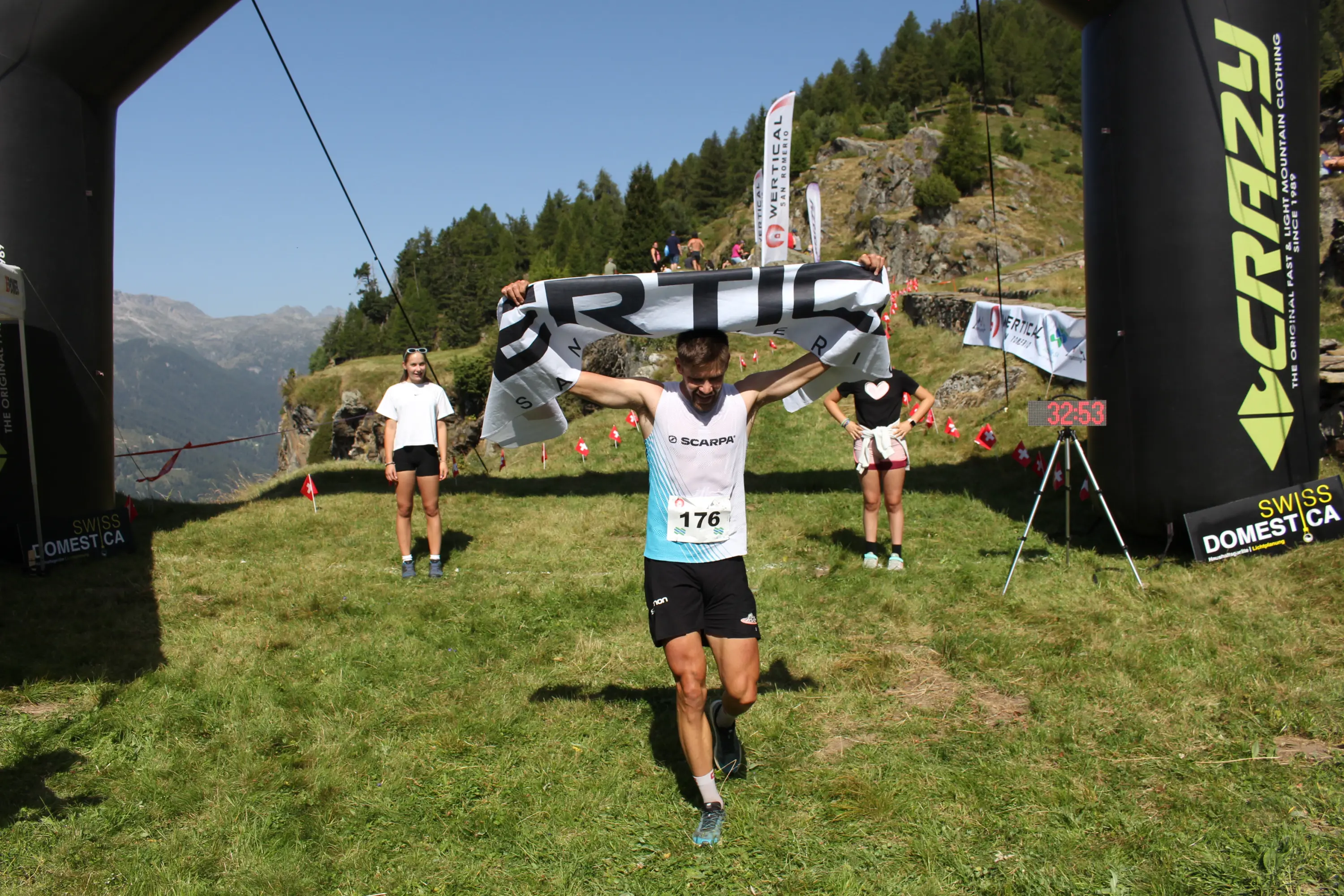 Un corridore taglia il traguardo sollevando un banner con la scritta "ARTIC". Sullo sfondo si vedono montagne verdi e persone che fanno il tifo.
