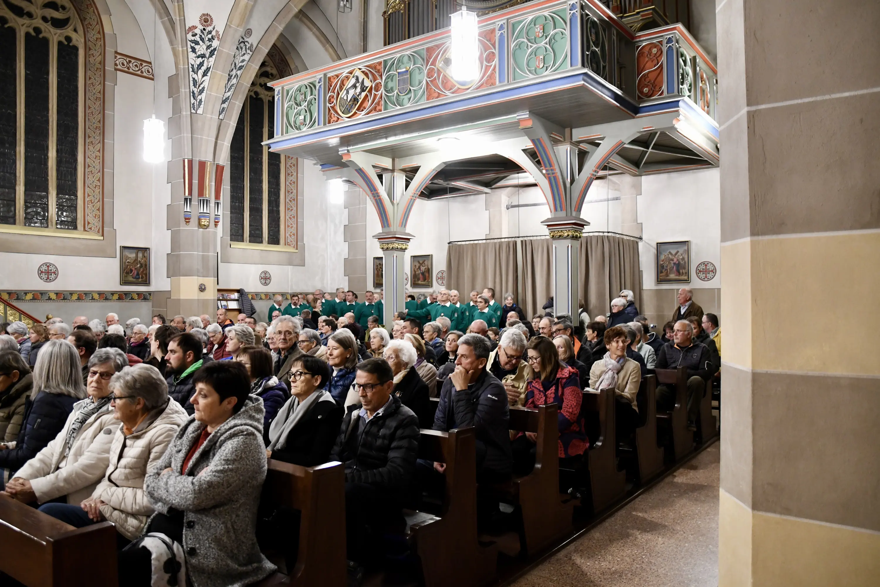 Una chiesa affollata con persone sedute sui banchi. Sullo sfondo, un gruppo di persone in abiti verdi è schierato. Le pareti sono decorate con vetrate e affreschi. L’atmosfera è solenne.