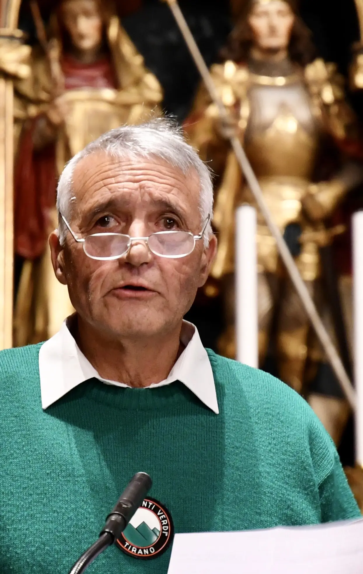 Uomo con capelli grigi e occhiali legge un discorso, indossando un maglione verde. Alle spalle, si possono vedere statue di figure storiche in un ambiente decorato.