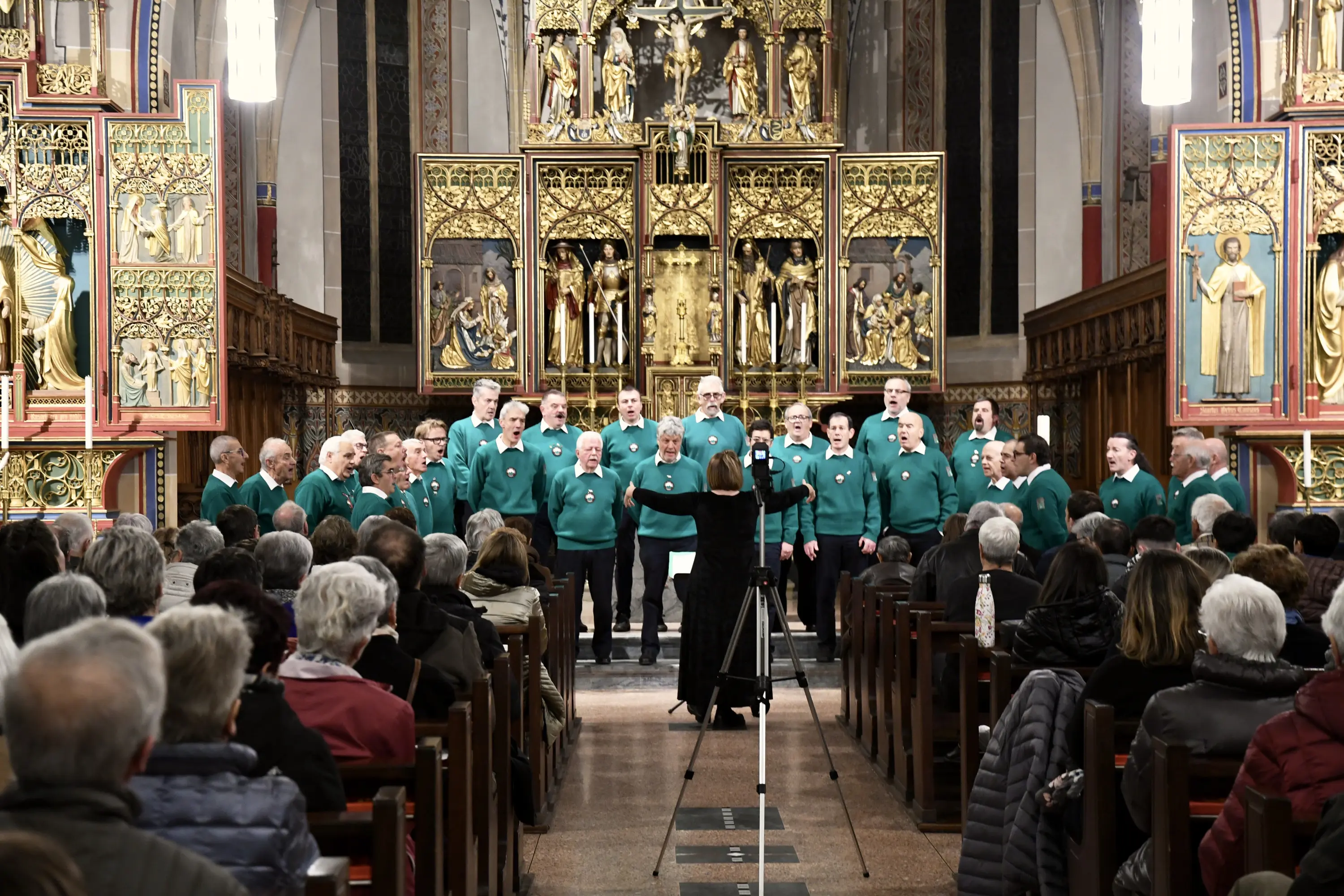 Un coro maschile in maglioni verdi si esibisce in una chiesa, davanti a un pubblico attento. Alle spalle del coro, un altare riccamente decorato aggiunge bellezza all'ambiente. Una direttrice guida la performance.