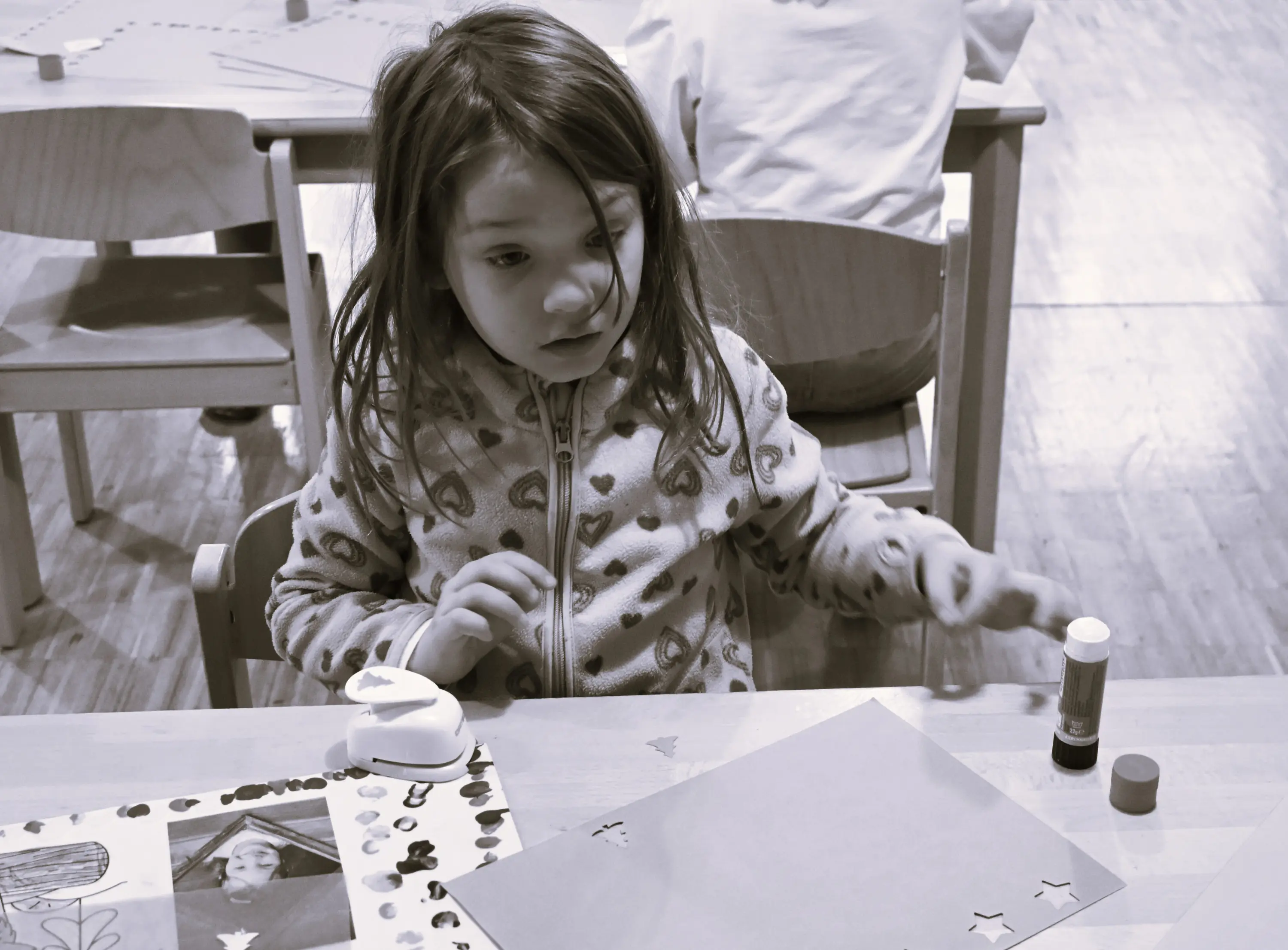 Una bambina, seduta a un tavolo, sta lavorando su un progetto artistico. Ha i capelli lunghi e mossi e indossa un pigiama con cuori. Sul