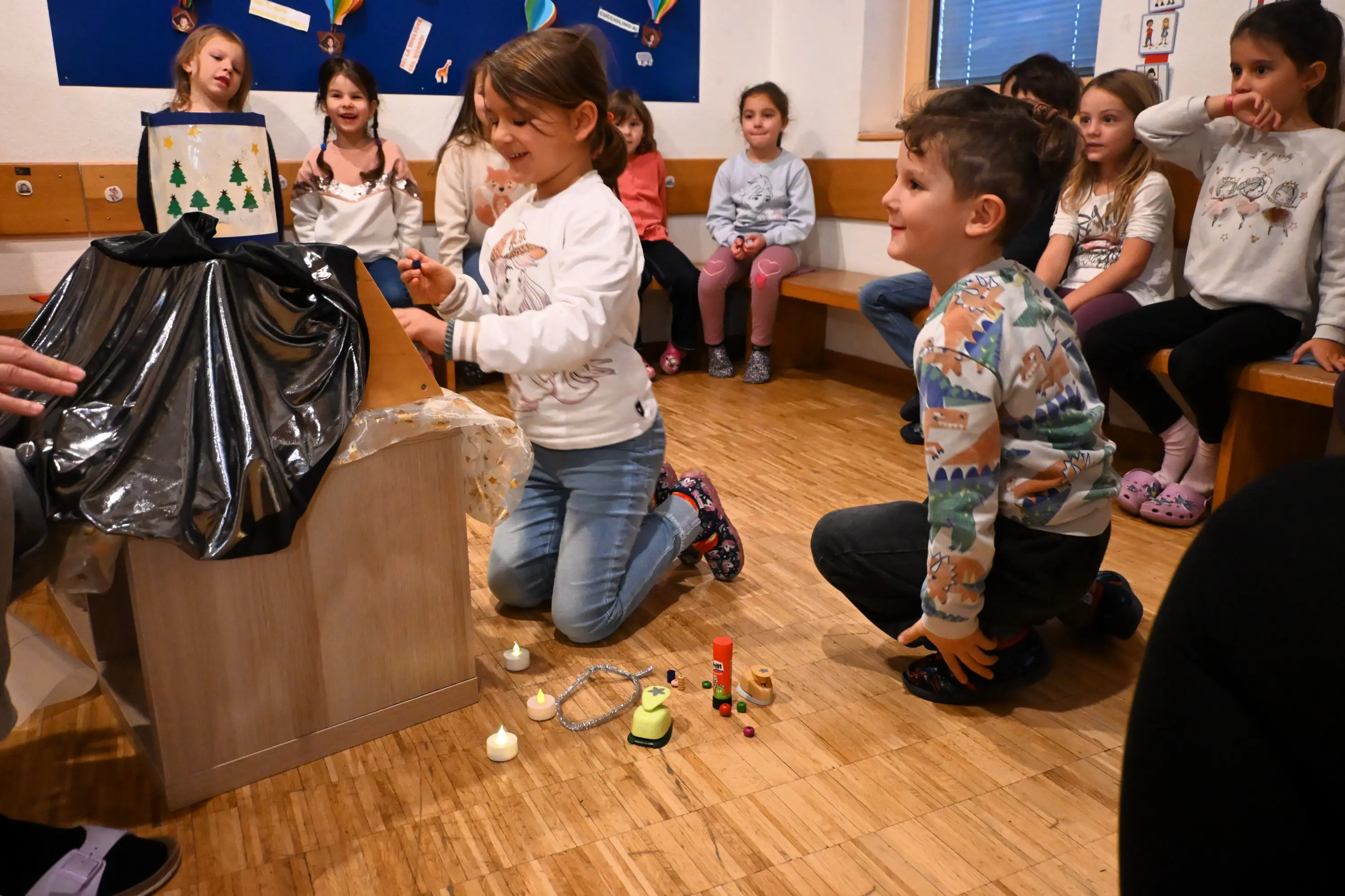 Due bambini stanno giocando a un tavolo, con diversi oggetti creativi disposti sul pavimento. Dietro di loro, un gruppo di bambini assiste, sorridendo e manifestando