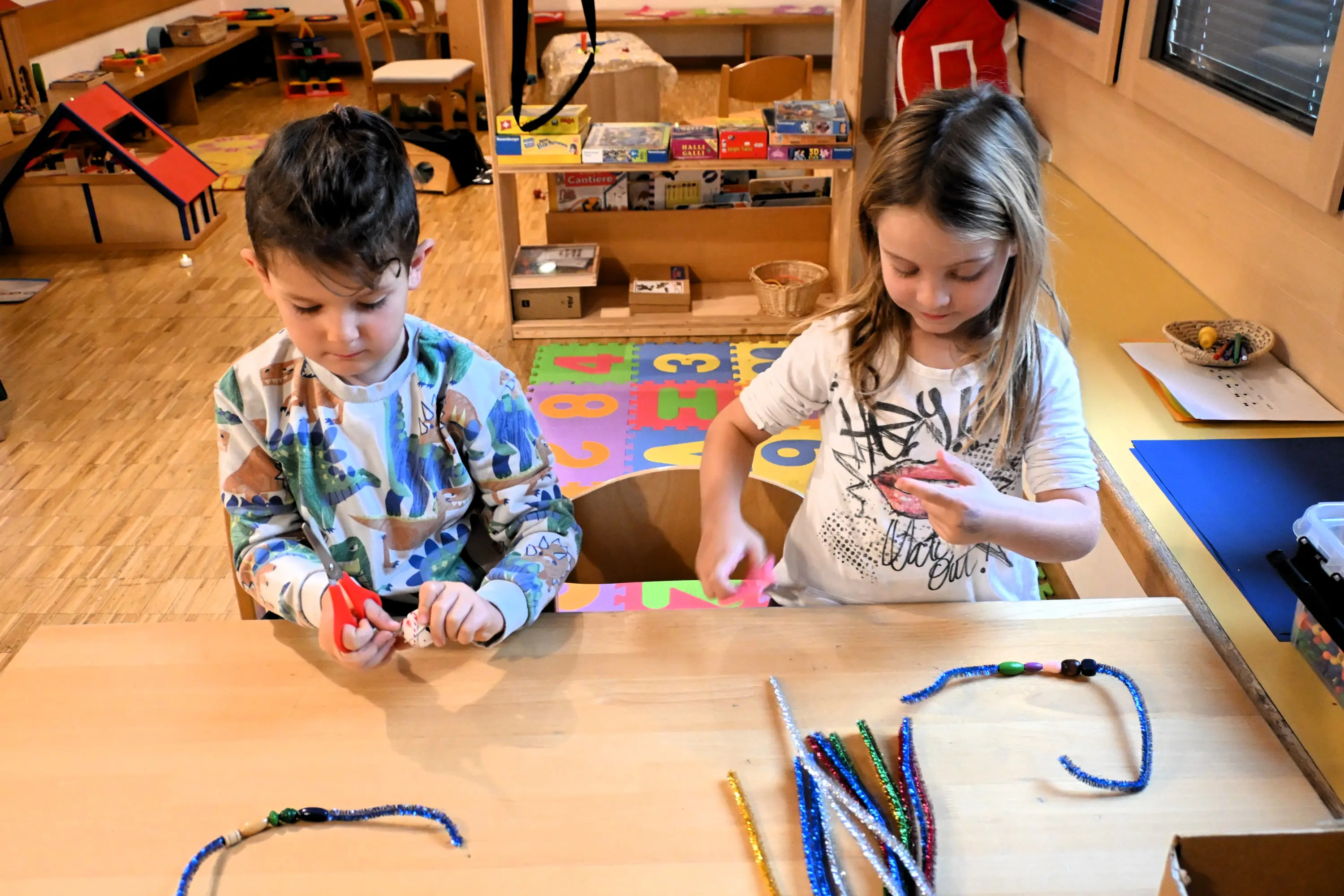 Due bambini stanno giocando a un tavolo, concentrati nell'attività di creazione. Il bambino a sinistra tiene un oggetto rosso, mentre la bambina a destra manipola