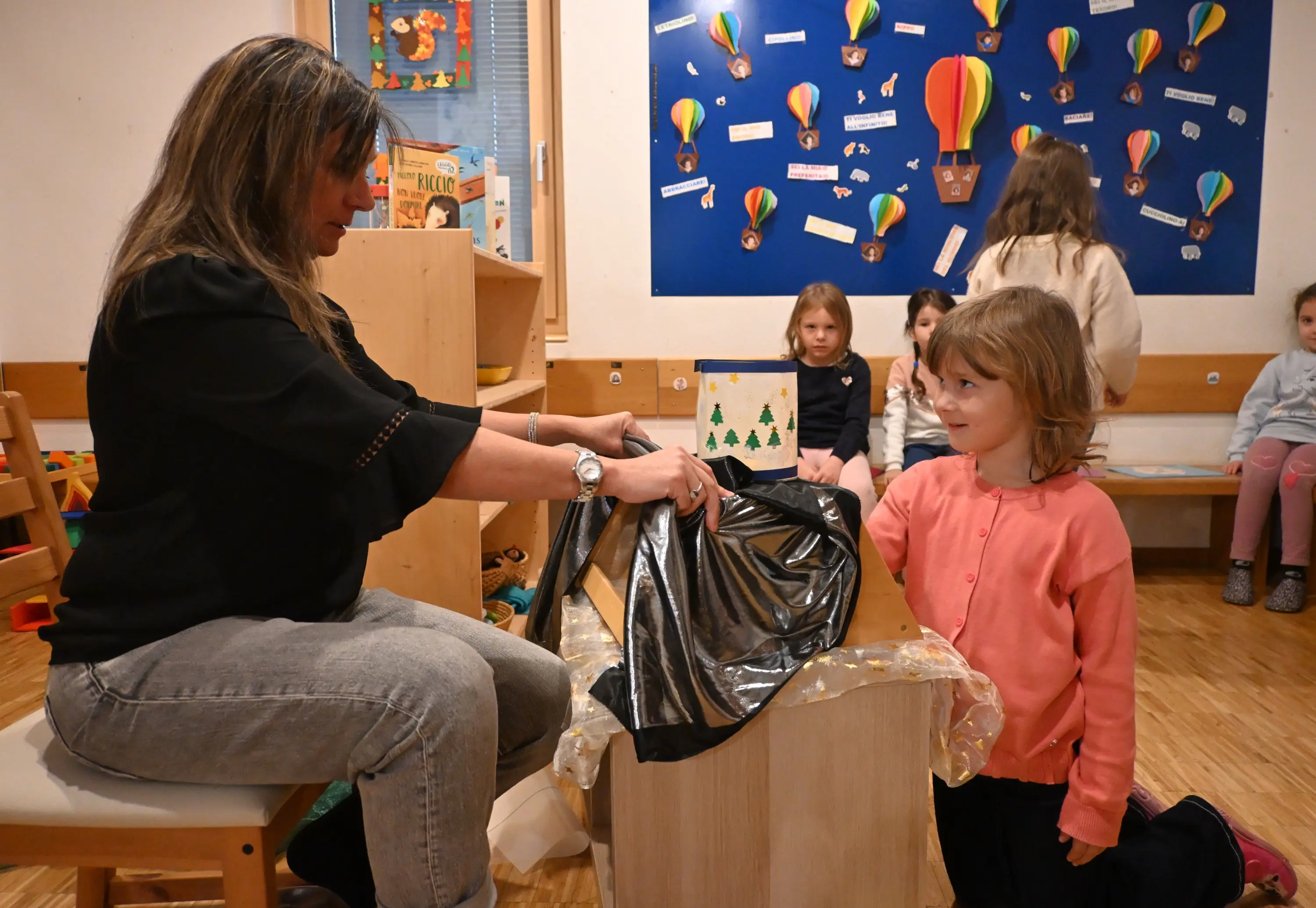 Una donna seduta apre un sacco nero con una bambina che osserva curiosa. Sullo sfondo, altri bambini sono seduti e guardano la scena in un'aula con decor