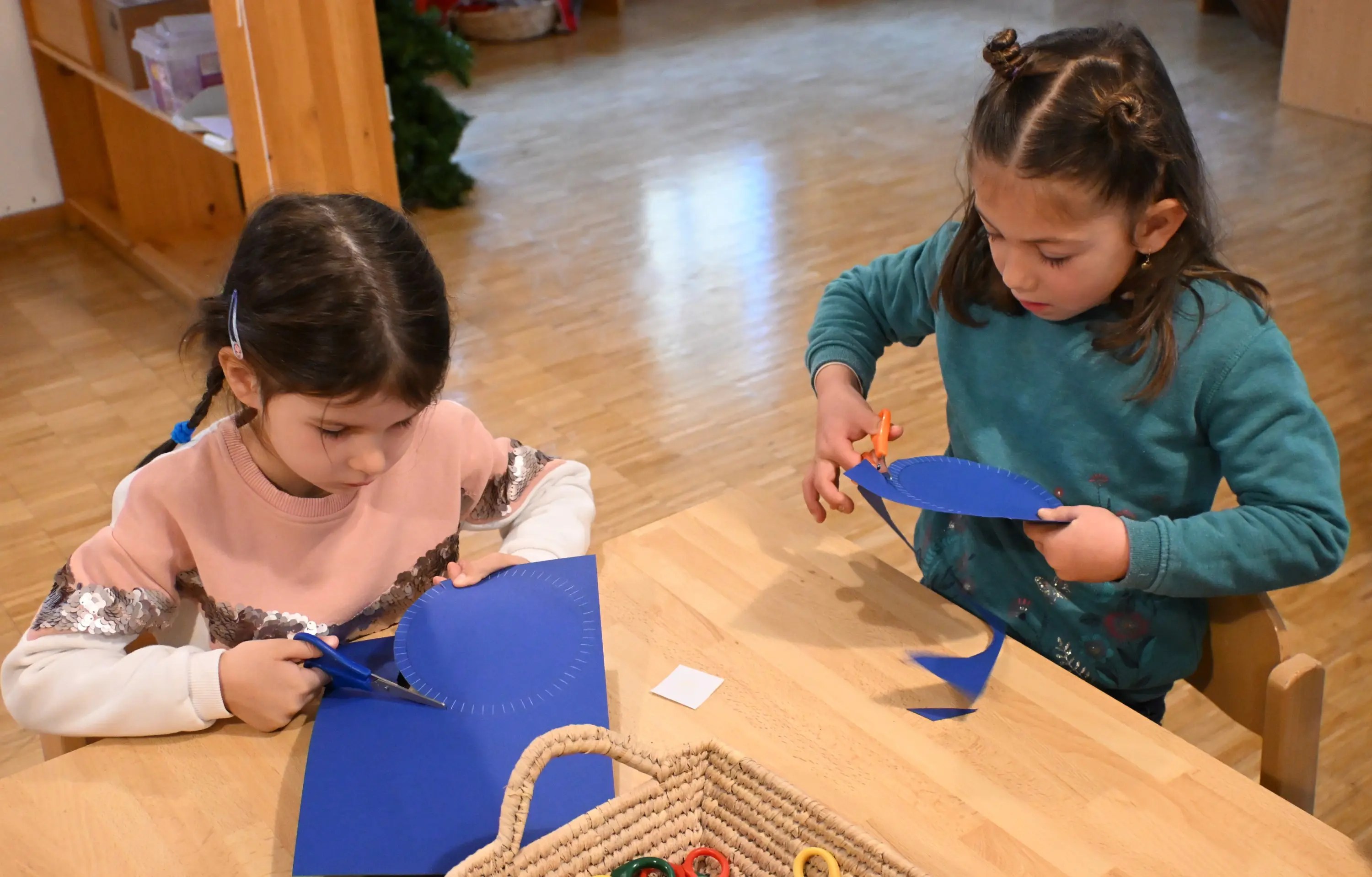 Due bambine sedute a un tavolo di legno, mentre stanno ritagliando forme blu con delle forbici. Una bambina indossa una maglietta chiara con dettagli lucc