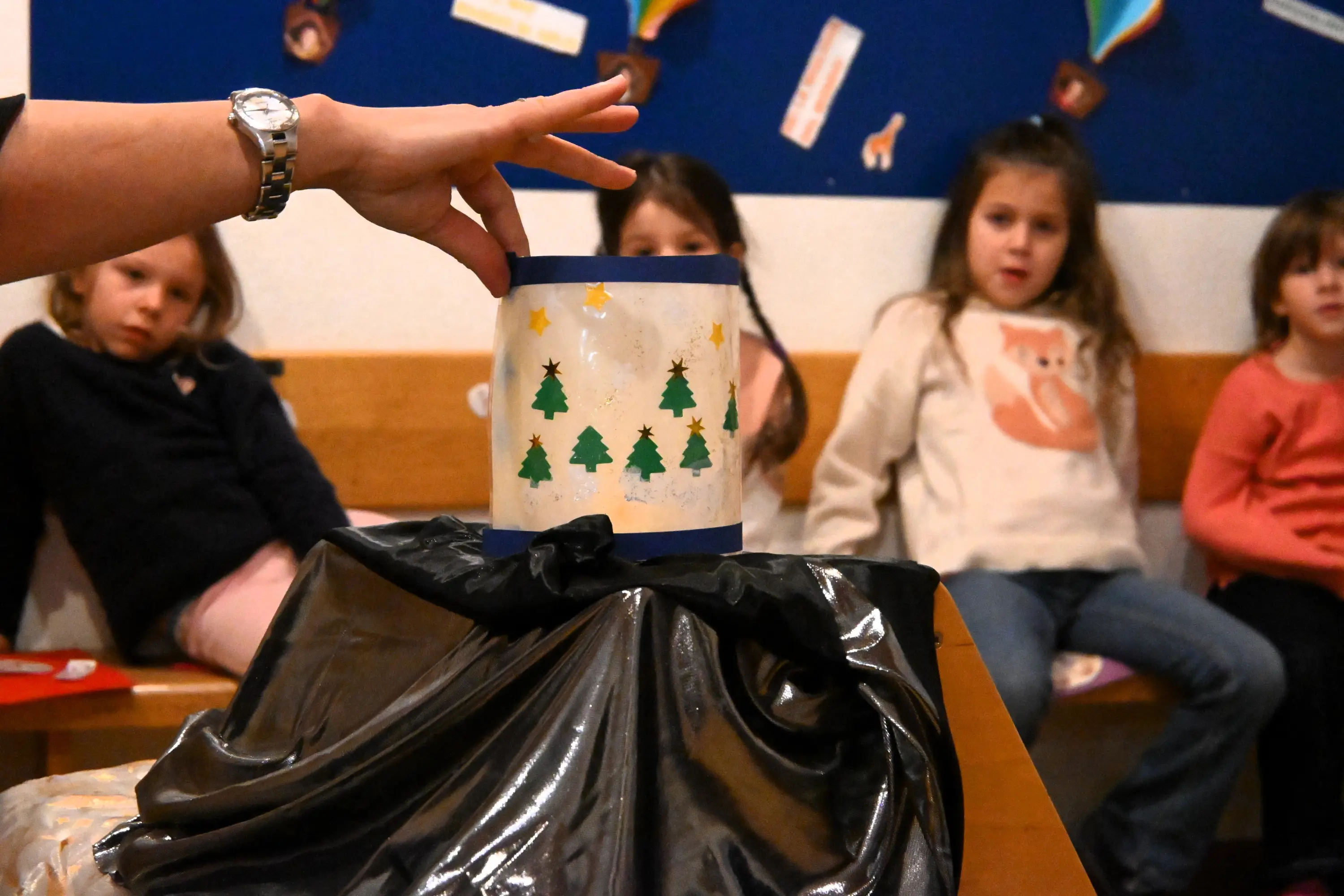 Una mano alza un barattolo decorato con alberi di Natale, mentre sullo sfondo tre bambini osservano attentamente. Su un tavolo è presente un drappo nero.