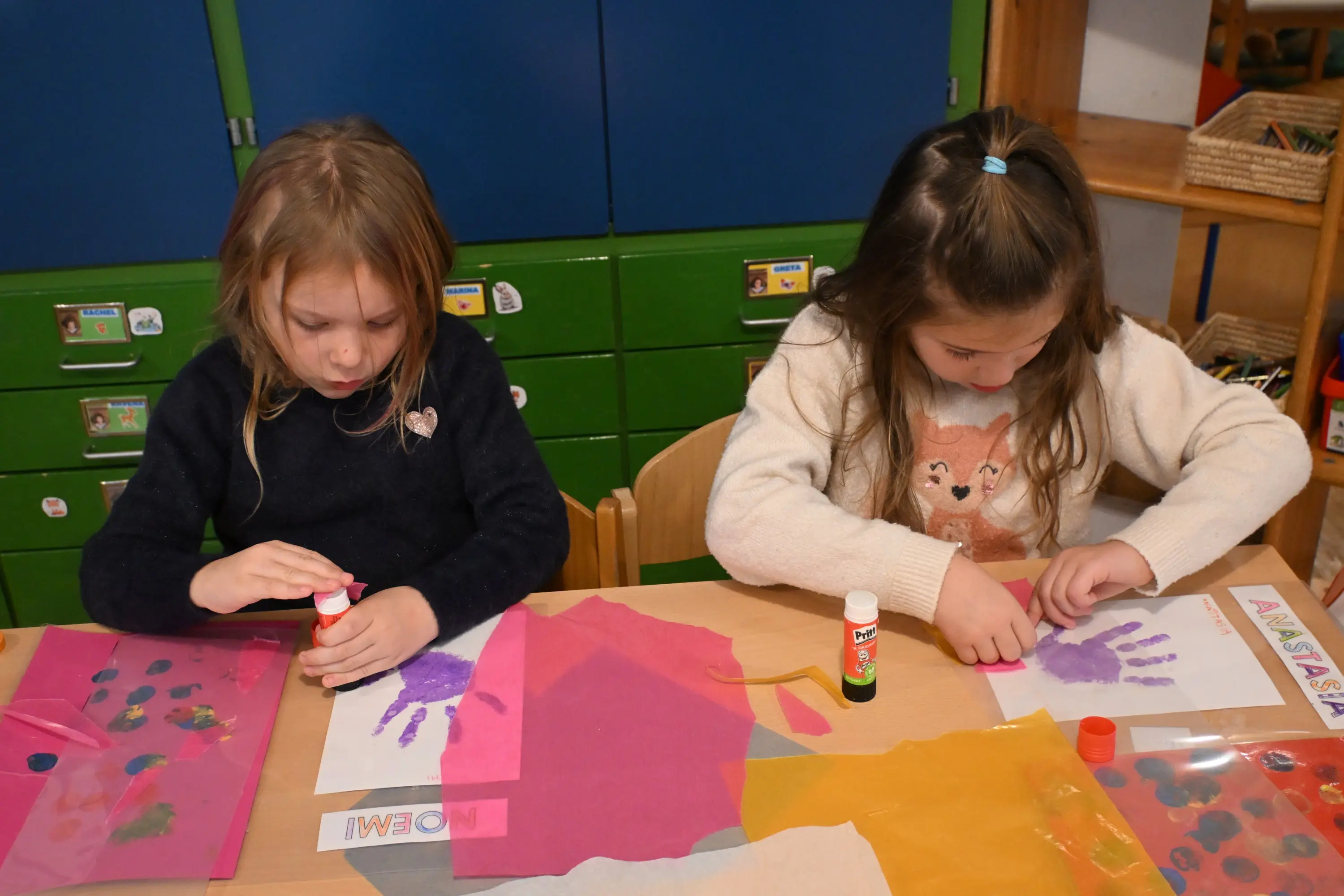 Due bambine sono sedute a un tavolo, mentre stanno creando dei progetti artistici con colla e carta colorata. Una delle bambine applica colla su una mano colorata