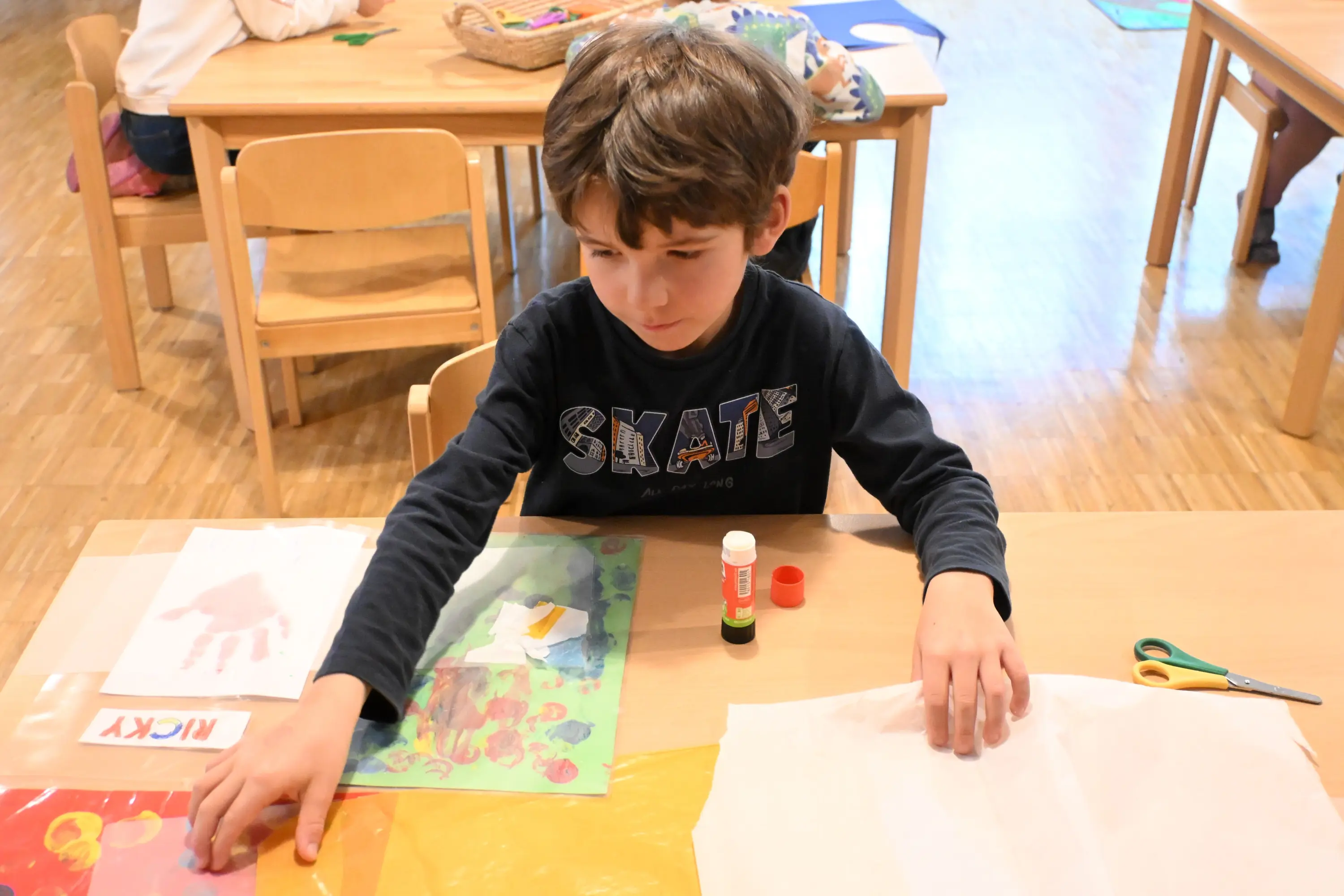 Un bambino seduto a un tavolo di legno mentre lavora a un progetto creativo. Davanti a lui ci sono vari fogli colorati, colla e forbici. Indossa una