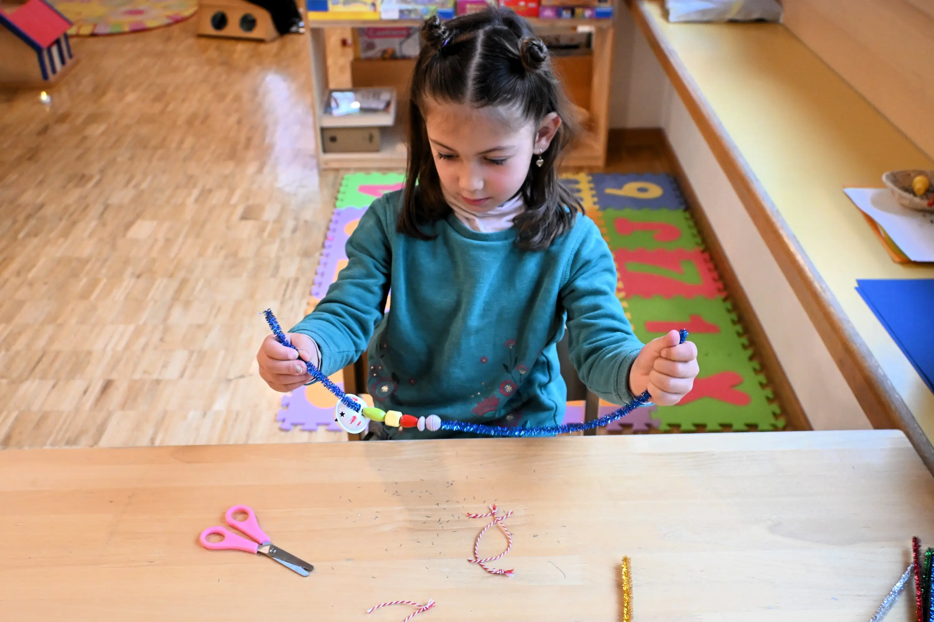 Una bambina seduta a un tavolo mentre crea un braccialetto con perline colorate e un filo blu. Ha i capelli scuri raccolti in due trecce e ind