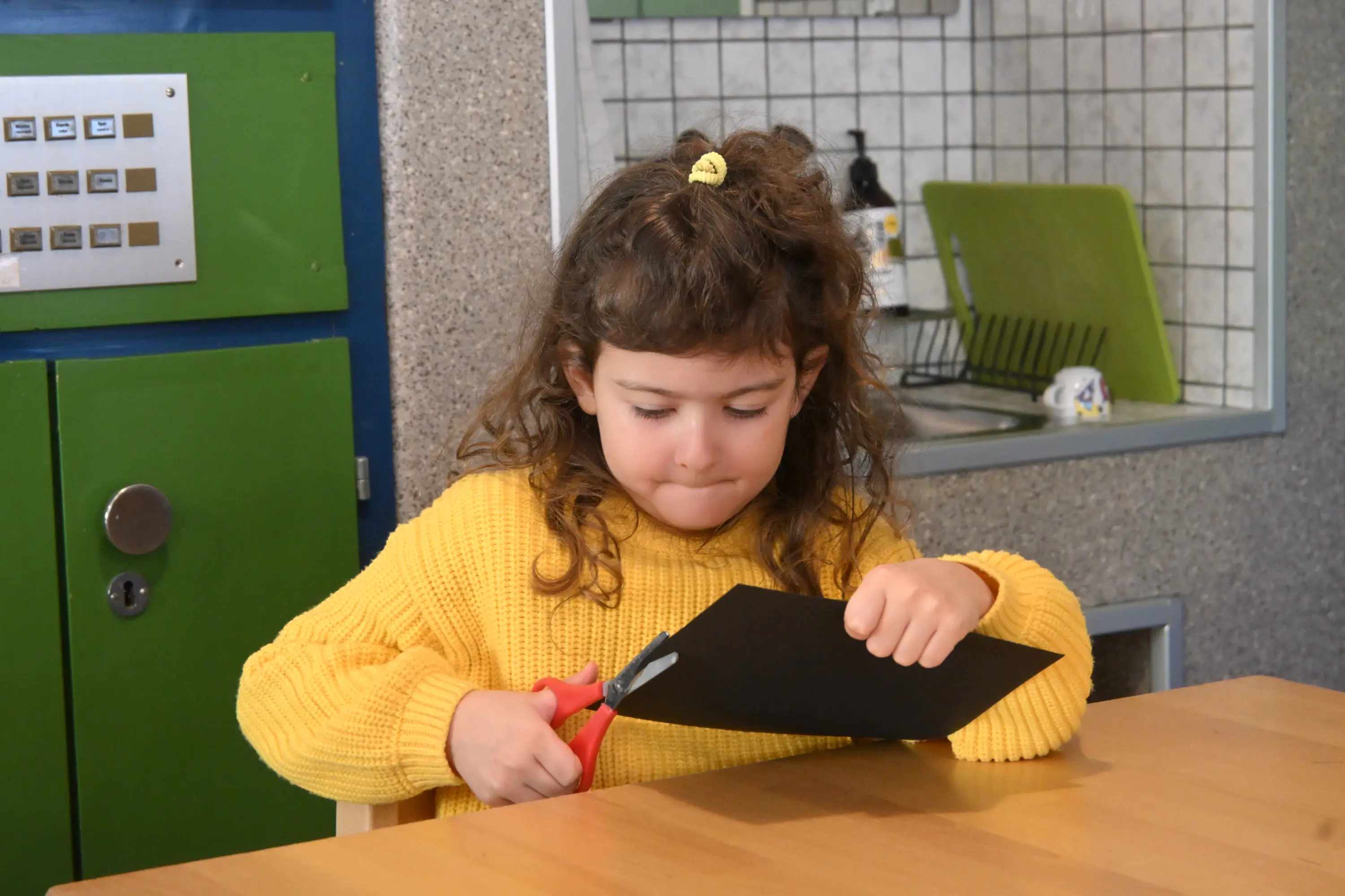 Una bambina con i capelli ricci e una maglia gialla sta tagliando un foglio di carta nera con delle forbici, concentrata al tavolo. Sullo sfondo