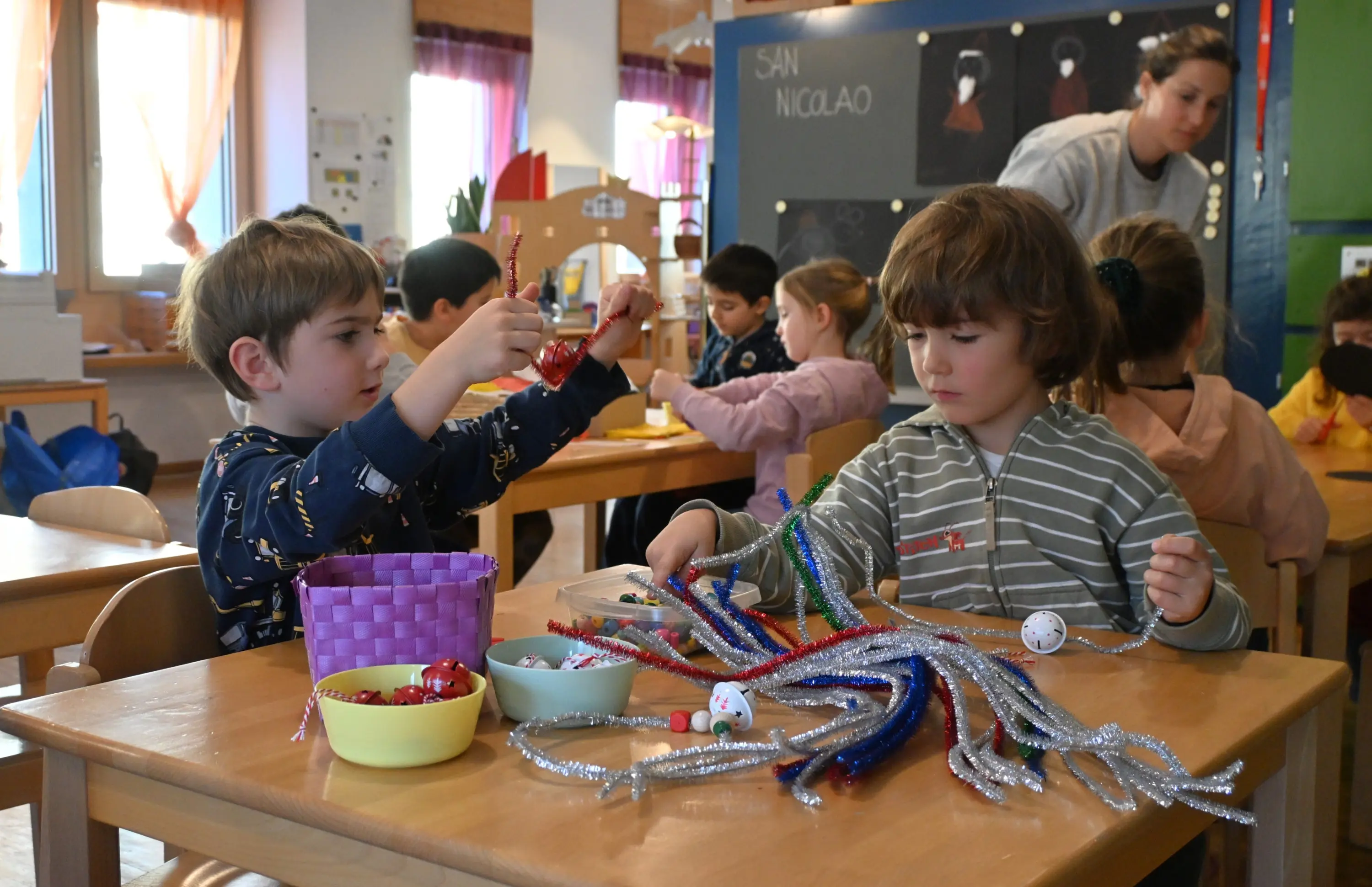 Due bambini seduti a un tavolo in una classe, intenti a creare oggetti con pipe cleaner colorati e decorazioni. Uno dei bambini tiene in mano un ornamento rosso mentre l