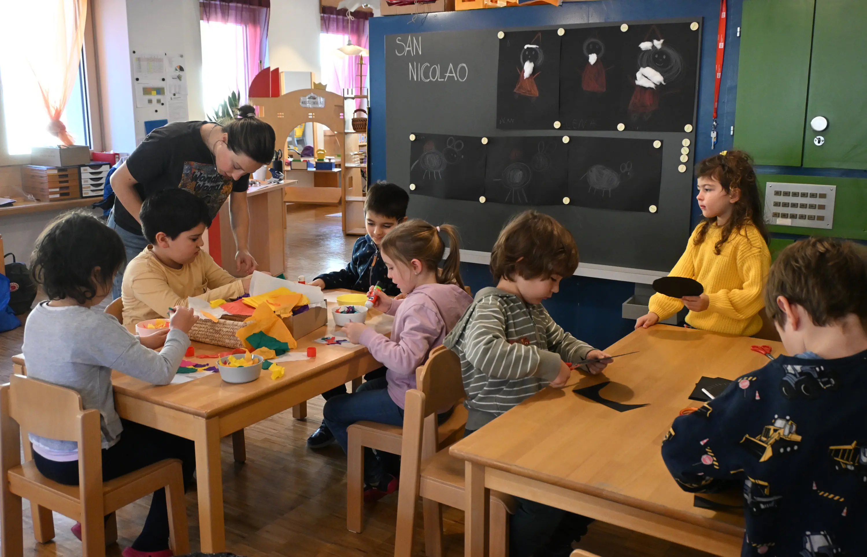 Gruppo di bambini seduti attorno a tavoli, impegnati in attività di arte e creatività con carta colorata e materiali vari. Un'insegnante assiste i bambini.
