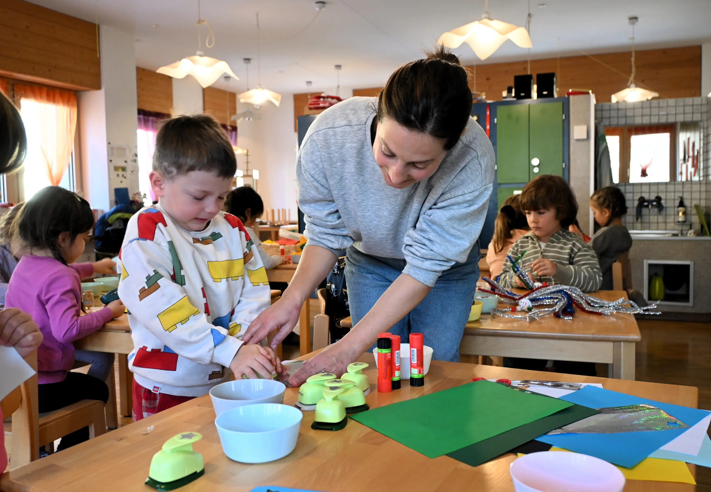 Un adulto aiuta un bambino a utilizzare della colla mentre altri bambini lavorano su progetti artistici a un tavolo. L'ambiente è luminoso e colorato, con materiali creativi