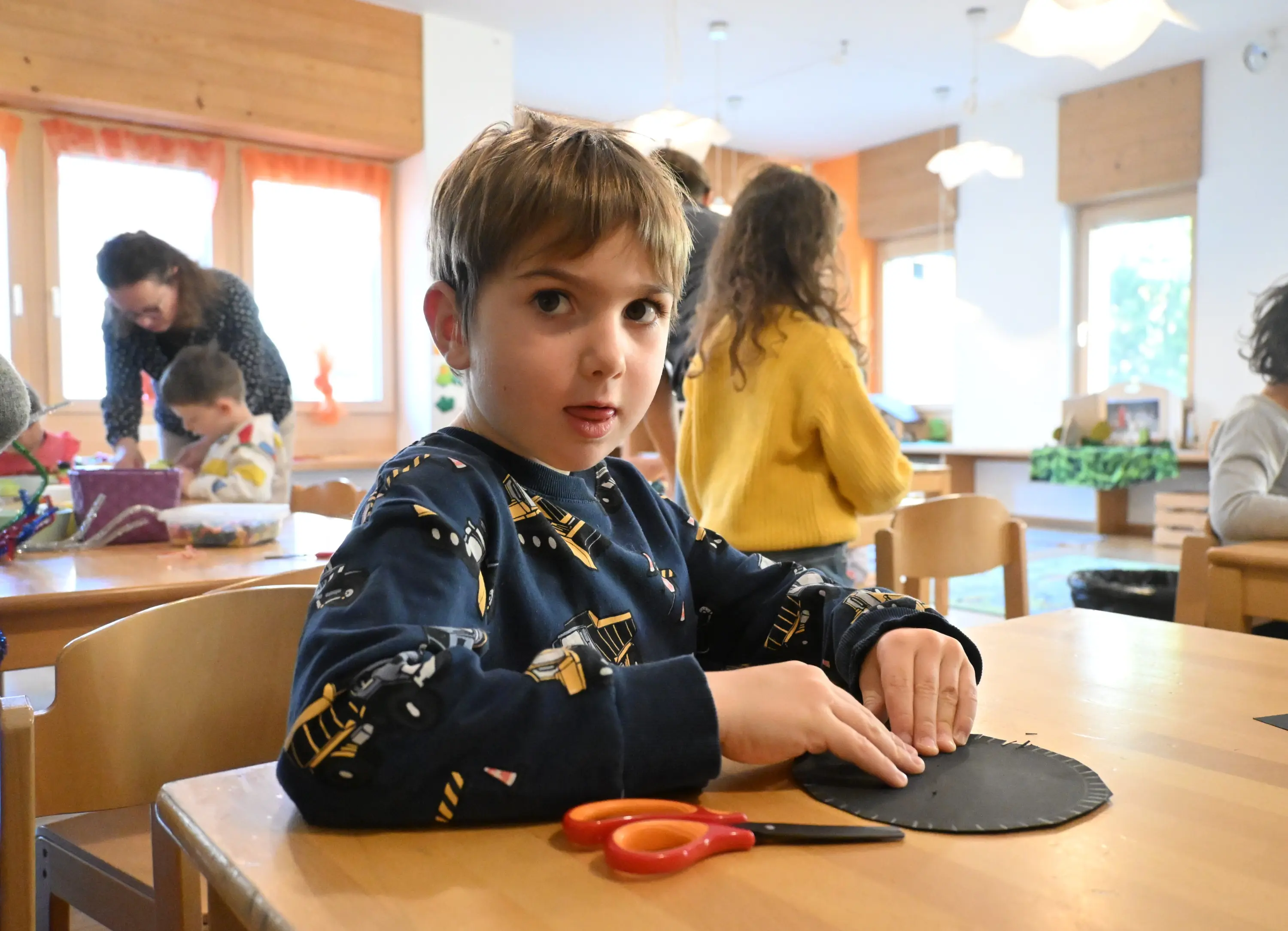 Un bambino con un maglione blu e motivi di camion è seduto a un tavolo, intento a tagliare un disco di carta con delle forbici. Sullo sfondo,