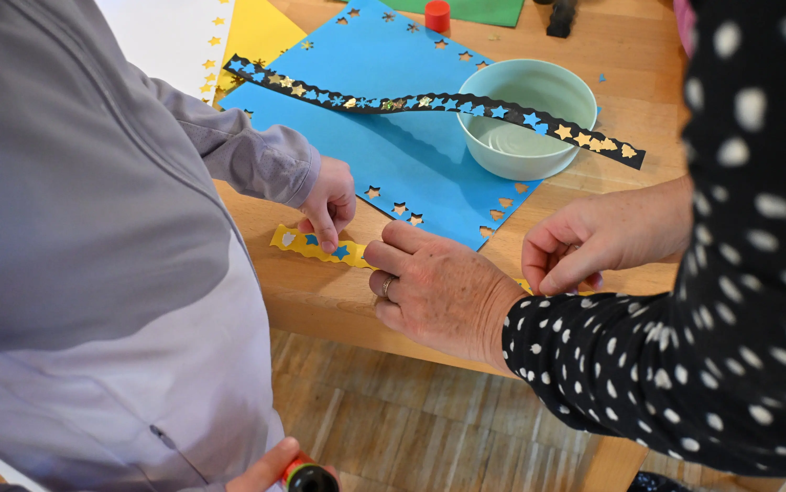 Bambini lavorano su un progetto creativo, applicando stelle di cartoncino colorato su un foglio blu. Una persona adulta assiste e aiuta.