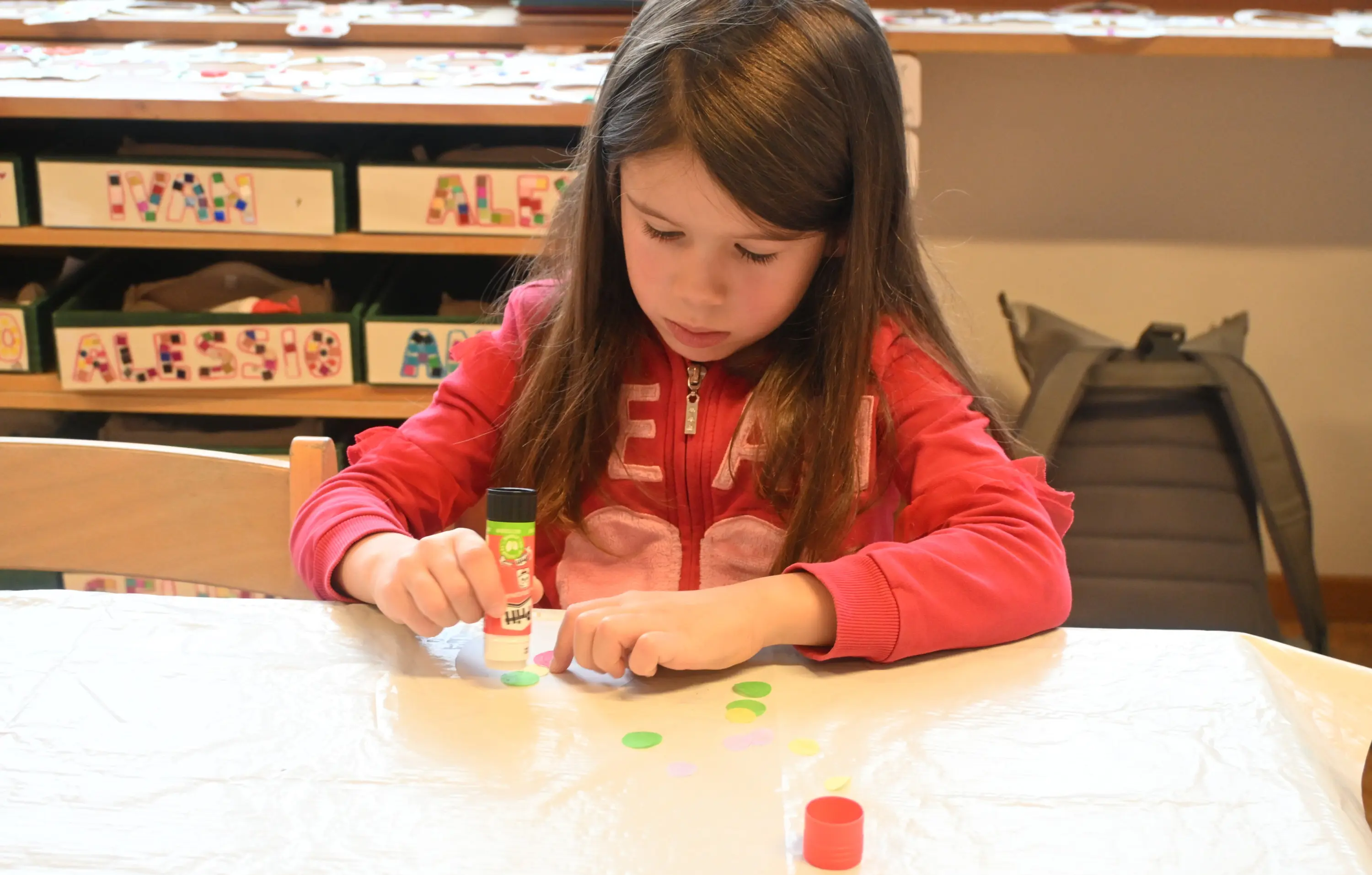 Una bambina sta incollando dei cerchi colorati su un tavolo. Indossa una felpa rossa e sembra concentrata nel suo lavoro creativo. Sullo sfondo ci sono materiali