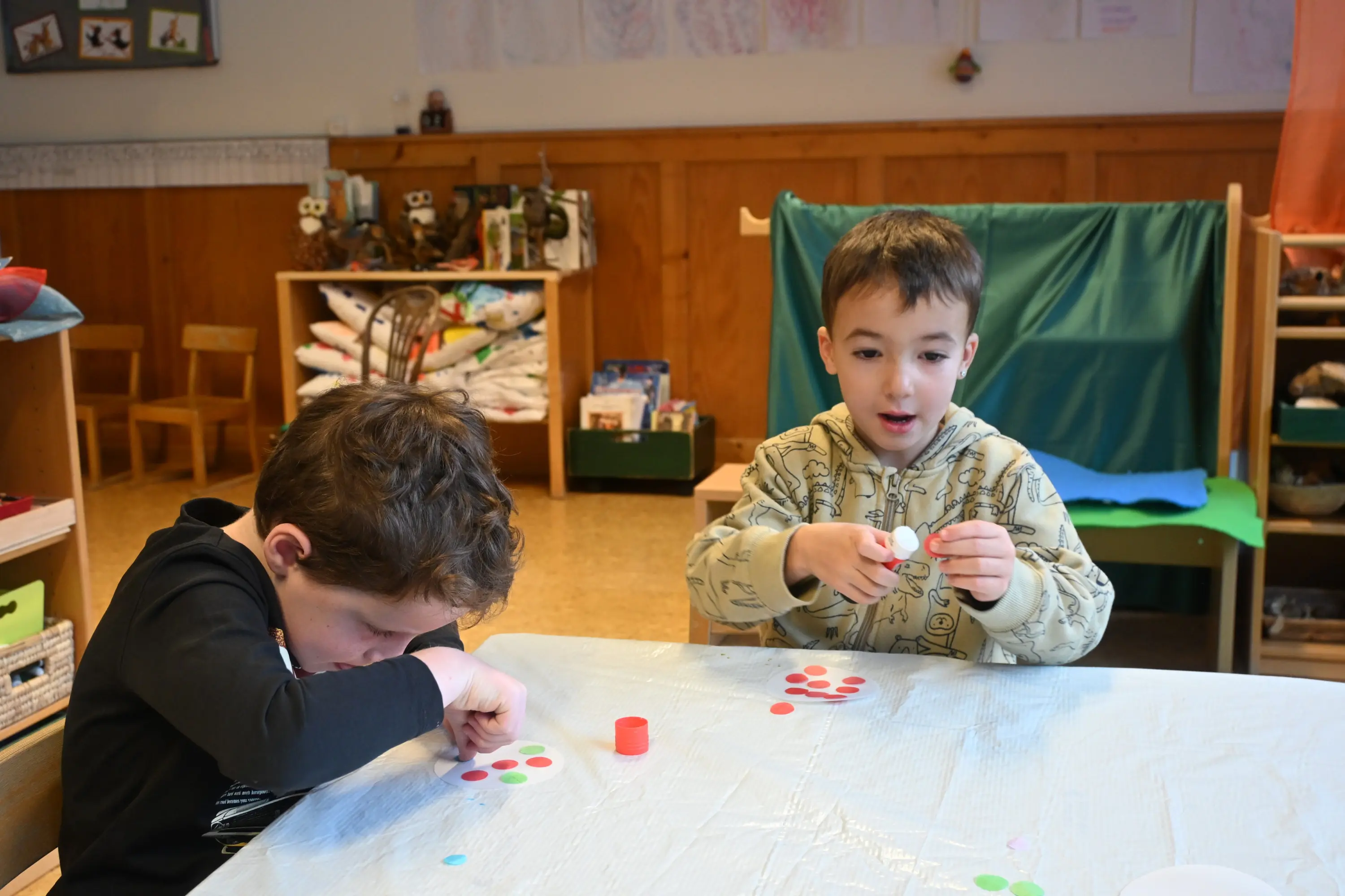 Due bambini stanno creando con dei colori e dei materiali sulla superficie di un tavolo. Uno dei bambini guarda attentamente mentre l'altro tiene un oggetto in mano, pronto a utilizzarlo. Sul