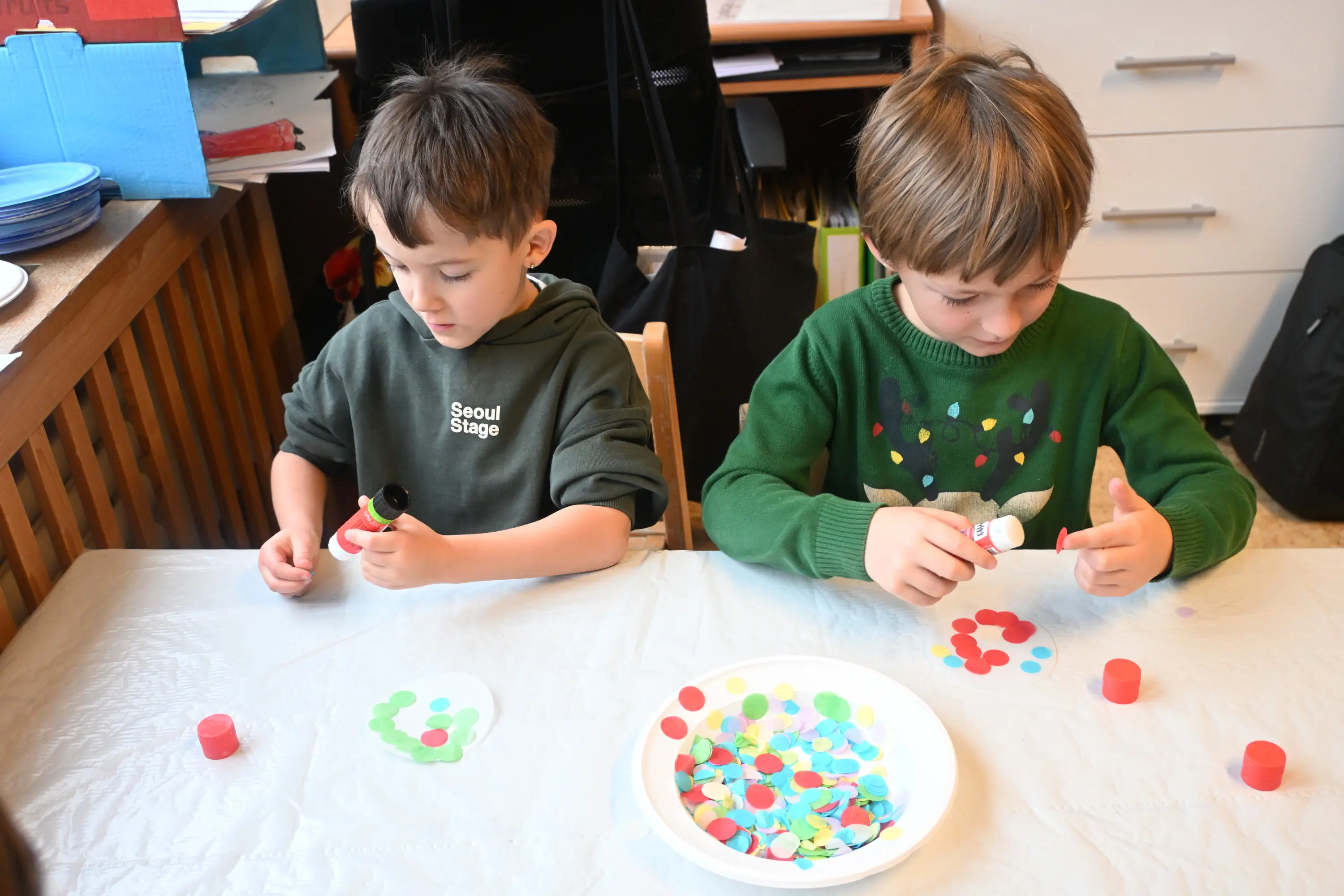 Due bambini seduti a un tavolo, intenti a incollare cerchi colorati su un foglio. Uno utilizza una colla, mentre l'altro tiene un tubetto di