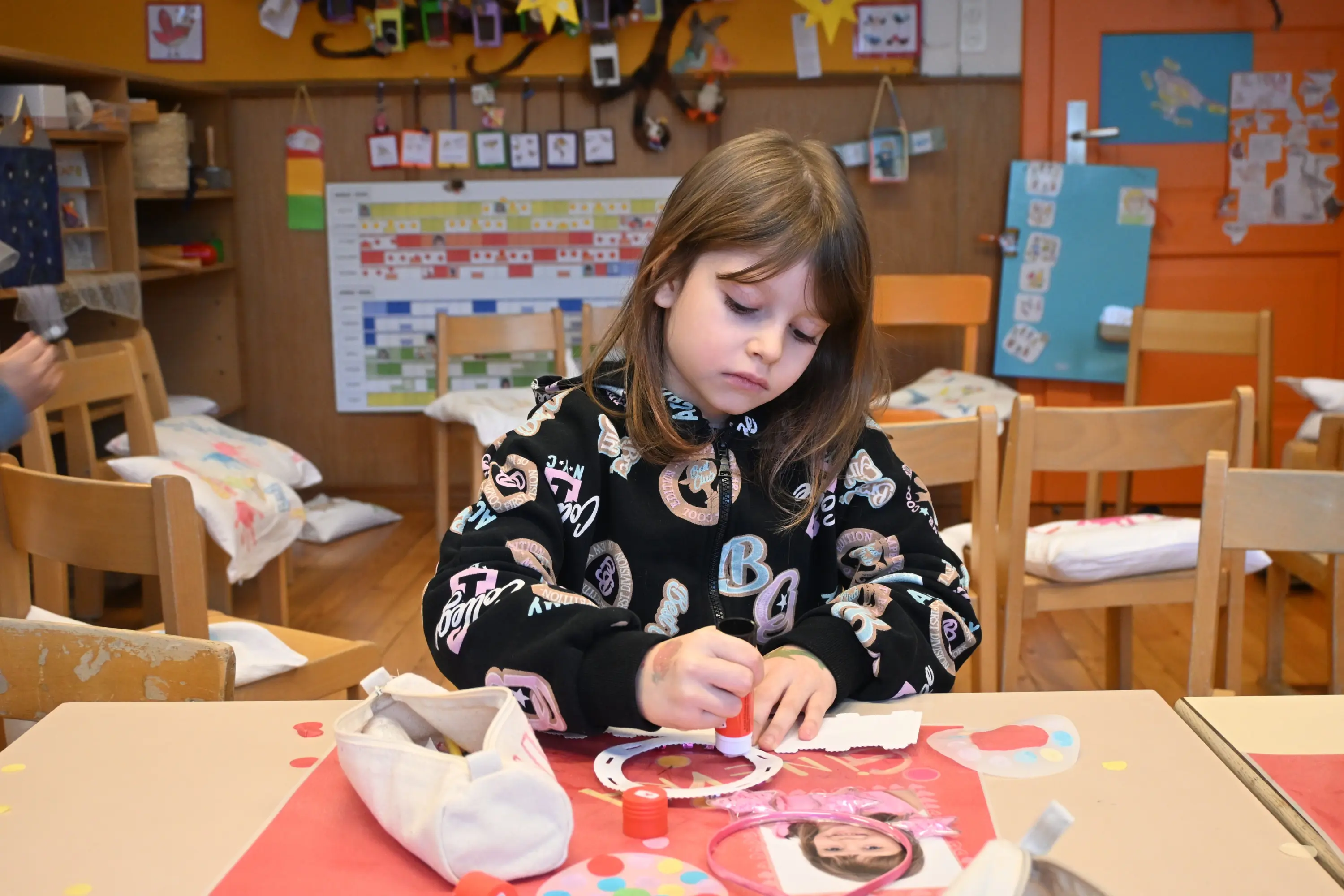 Una bambina è seduta a un tavolo in un'aula, mentre colora e lavora su un progetto artistico. Indossa una felpa nera con grafiche colorate
