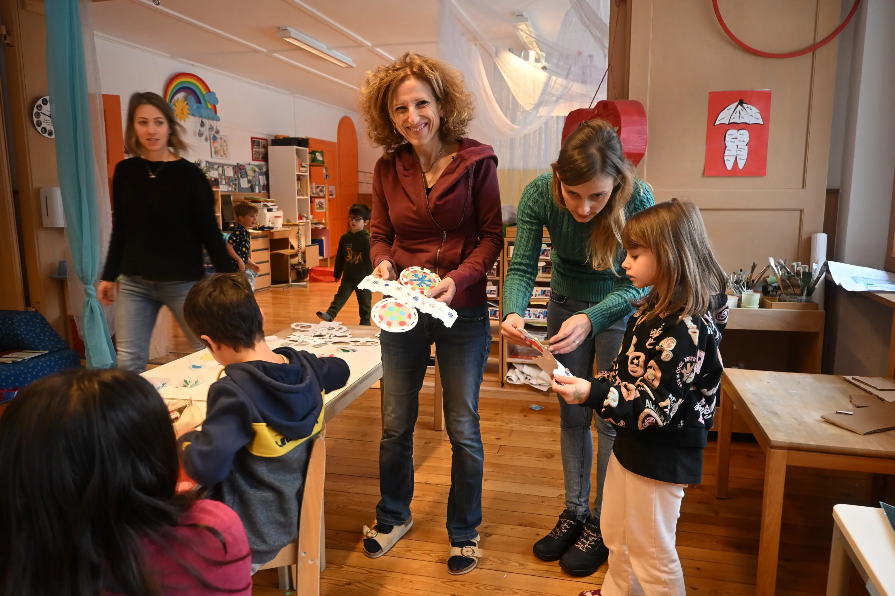 Un'aula creativa con bambini che partecipano a attività artistiche. Due adulti, una donna con i capelli ricci e un'altra con i capelli lisci, interagiscono con una