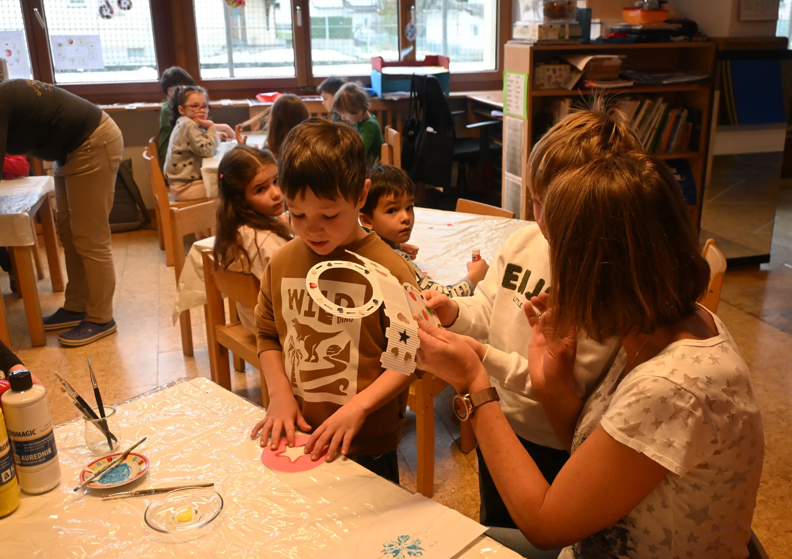 Un'insegnante mostra a un bambino come decorare un oggetto artigianale, circondati da altri bambini impegnati in attività creative in un'aula luminosa.