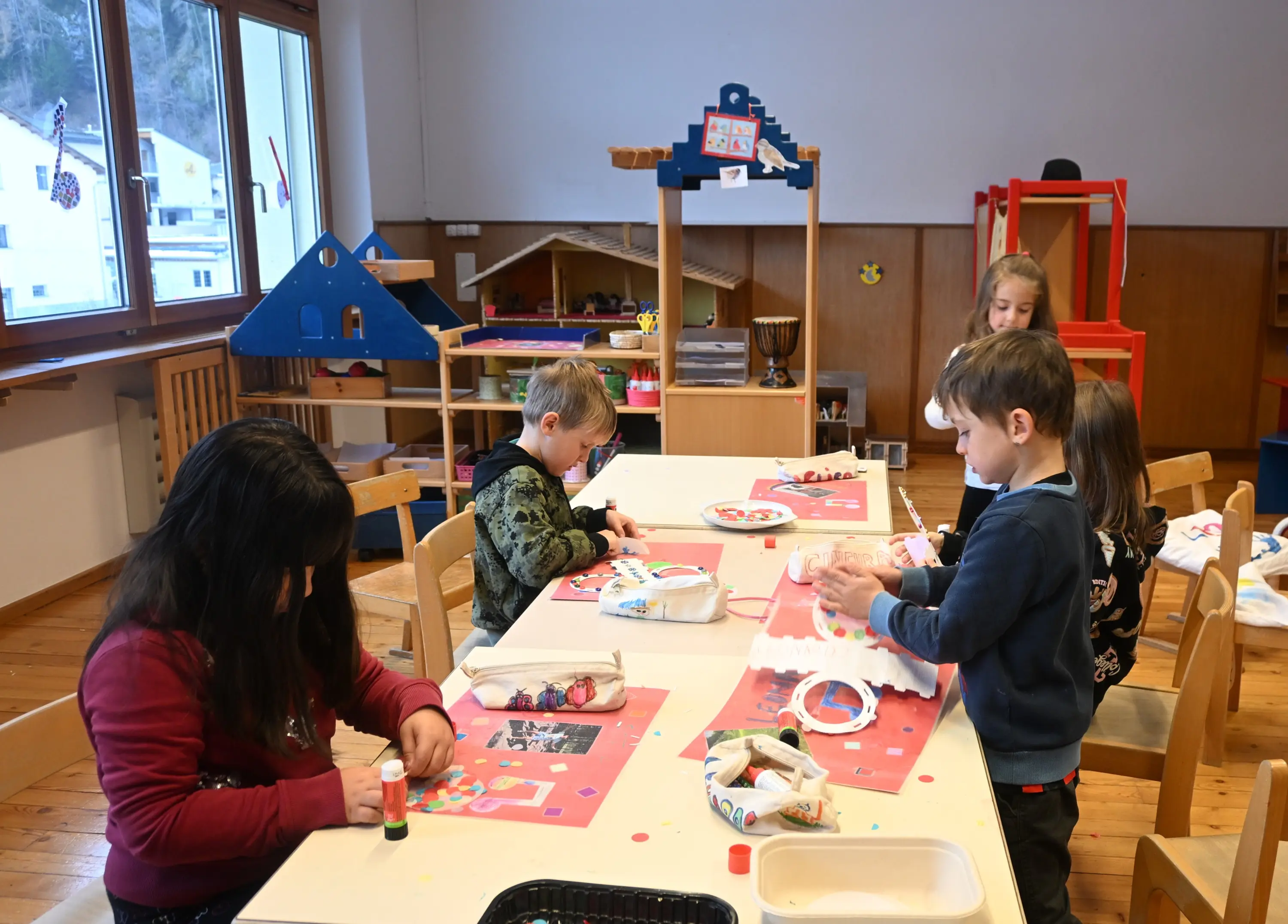 Bambini di diverse età lavorano a progetti artistici su tavoli in un'aula luminosa. Hanno materiali come carta colorata, colla e forbici e si concentr
