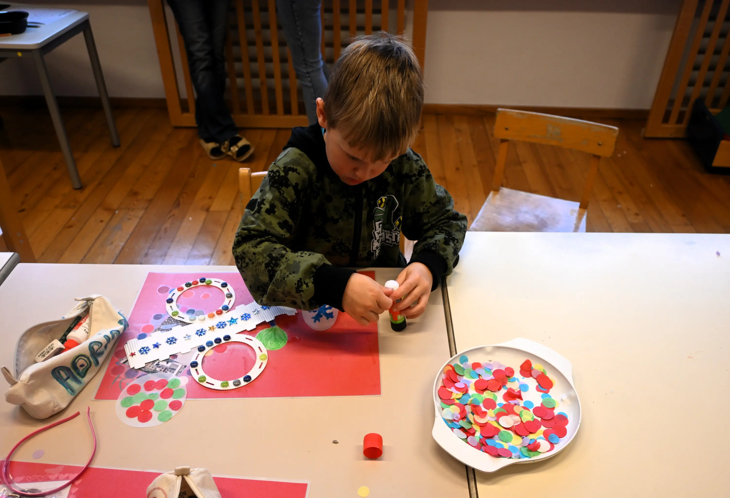 Bambino seduto a un tavolo, impegnato a incollare dei pezzi di carta colorata su un foglio rosso. Si vedono colori e materiali per il crafting